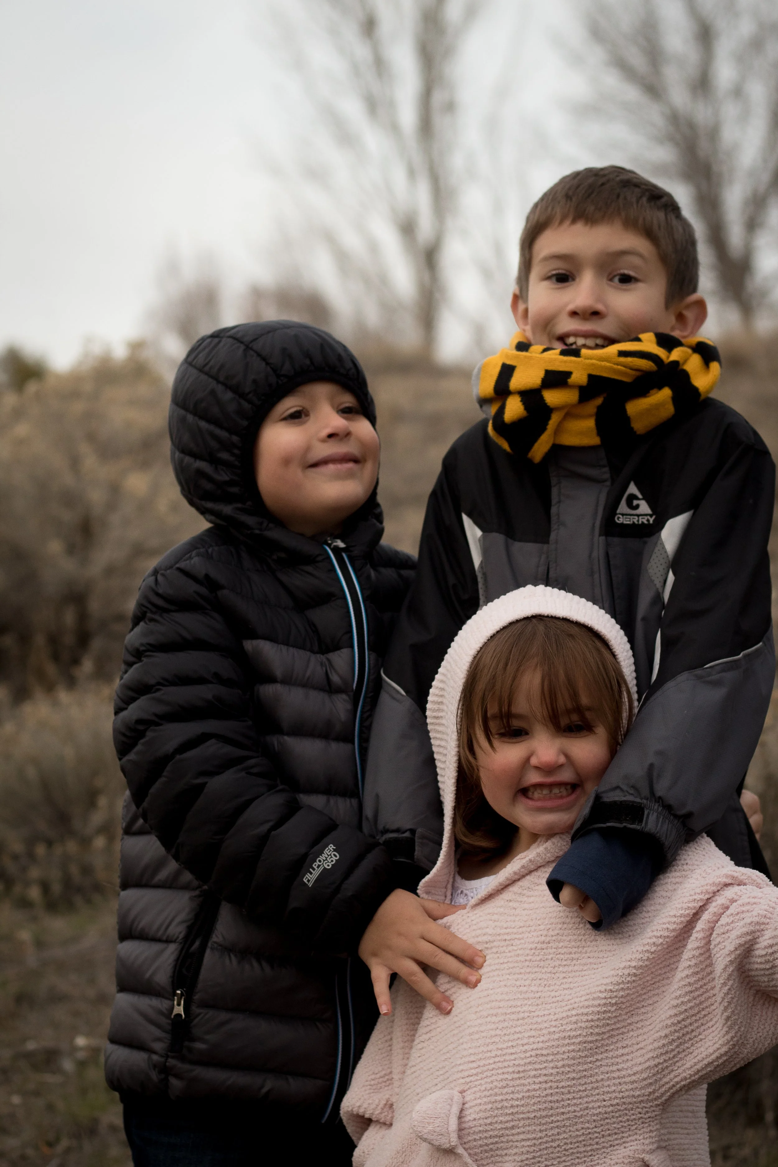 Three children outdoors, dressed in warm jackets and scarves, smiling and making funny faces.