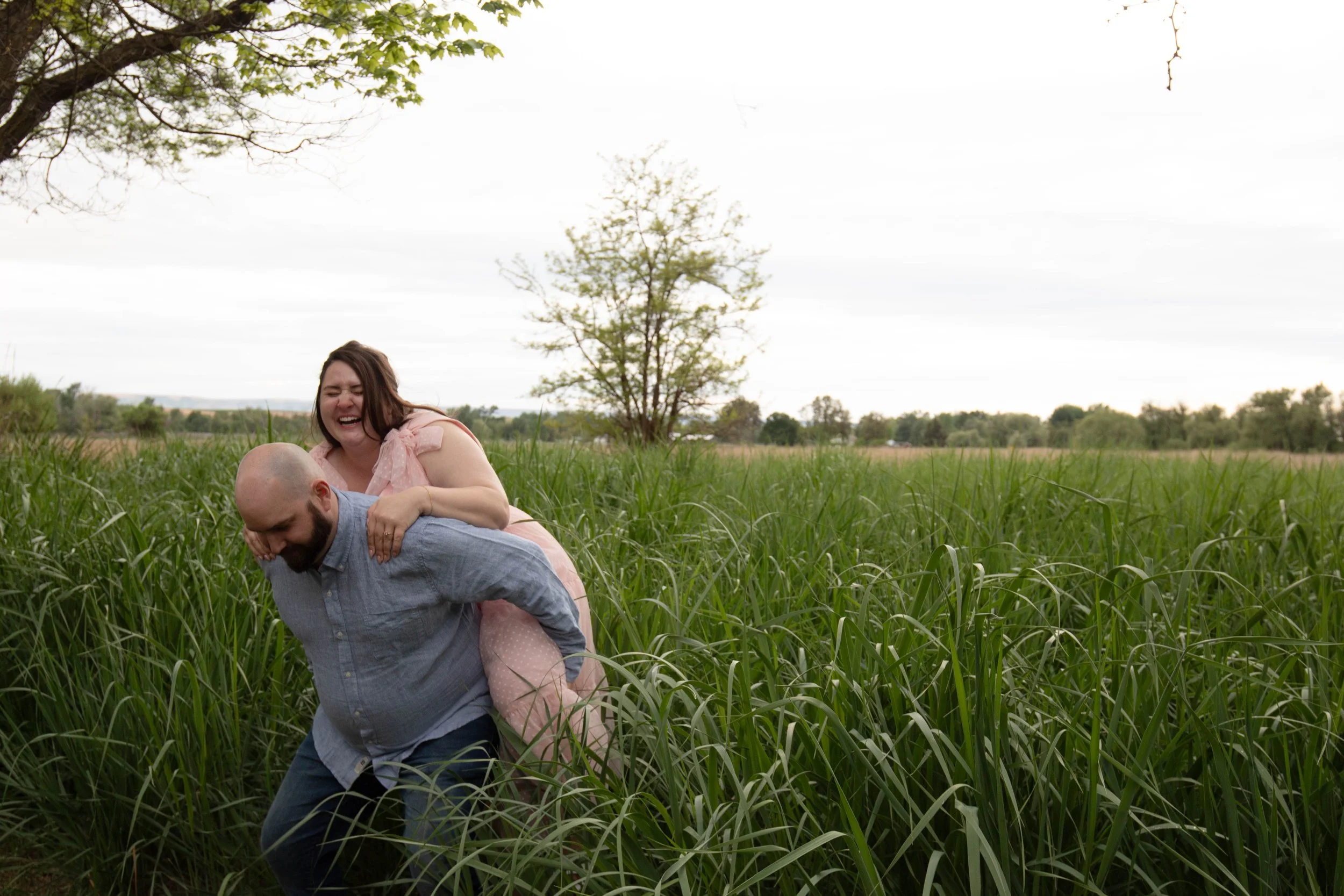A woman laughing and smiling playfully on a man’s back in a grassy field with a tree in the background.