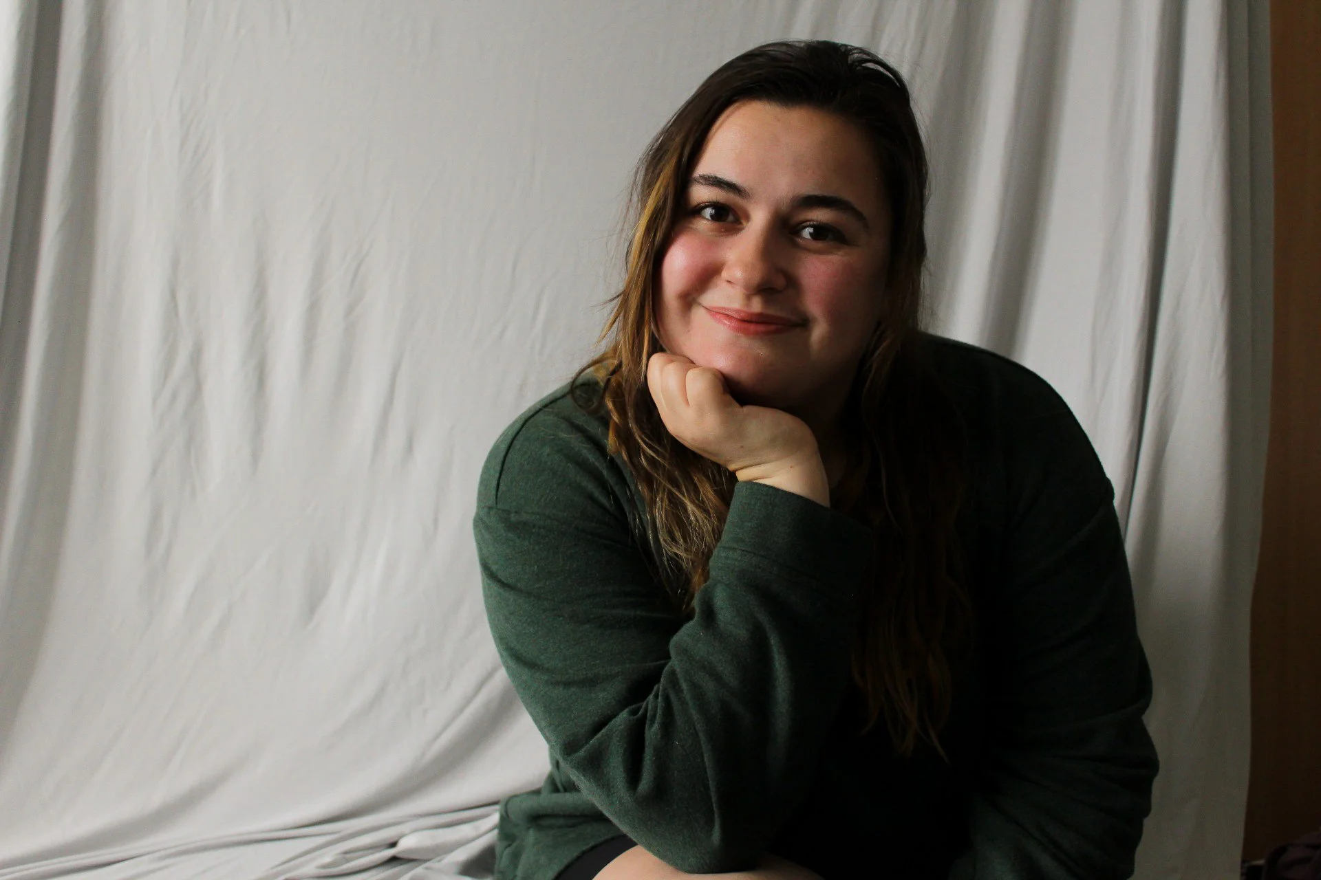 A woman with long brown hair, wearing a dark green sweatshirt, sitting in front of a light-colored backdrop, smiling and resting her chin on her hand.