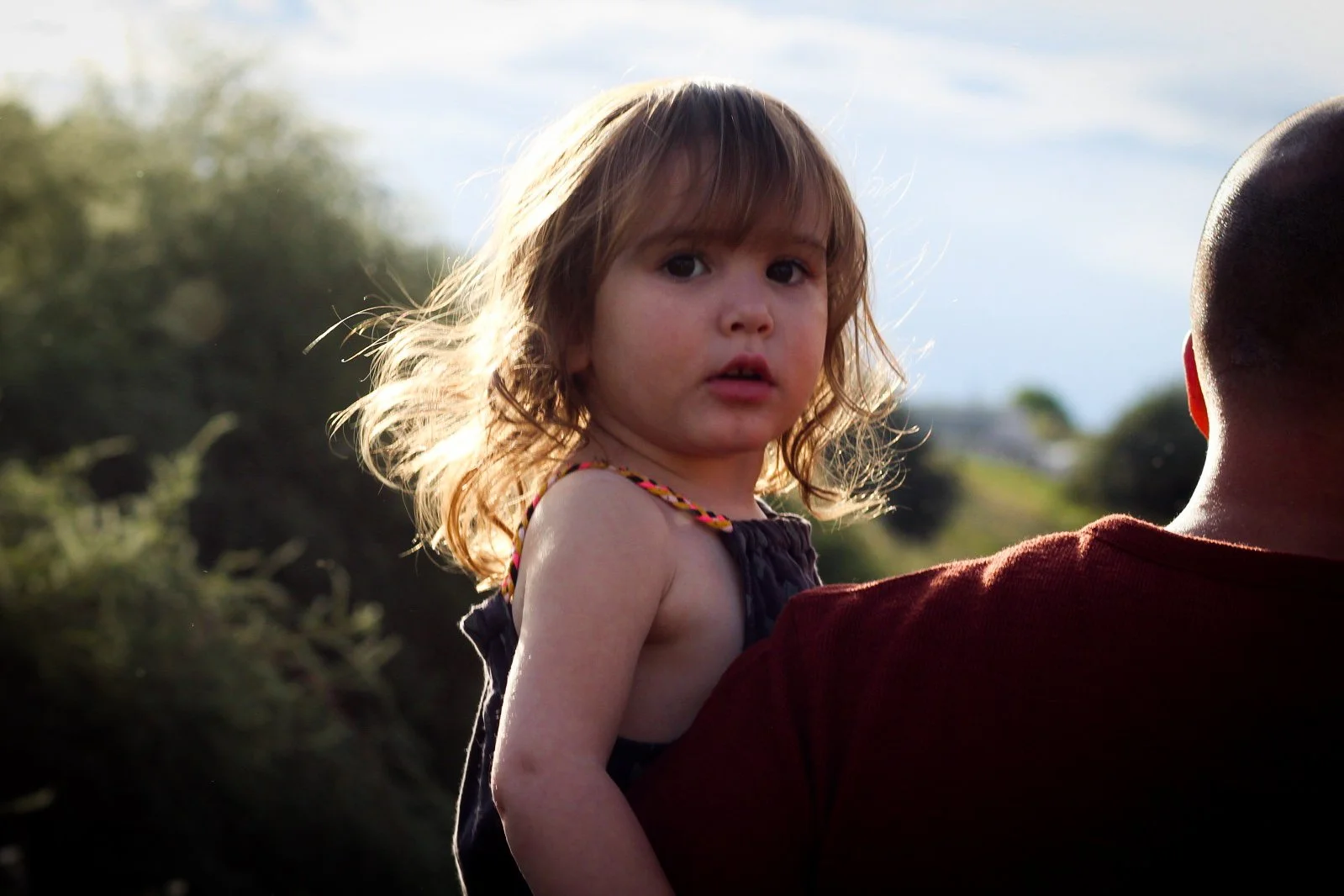Young girl with shoulder-length light brown hair and wearing a dark sleeveless top, being held by a person wearing a red shirt outdoors with a blurred natural background and a partly cloudy sky.