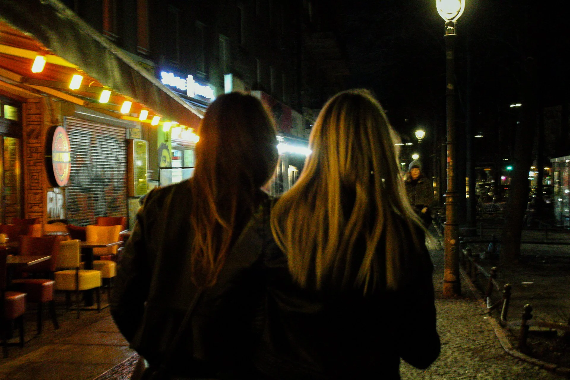 Two women with long hair walking on a city sidewalk at night, illuminated by streetlights and shop signs, near a restaurant or cafe with outdoor seating.