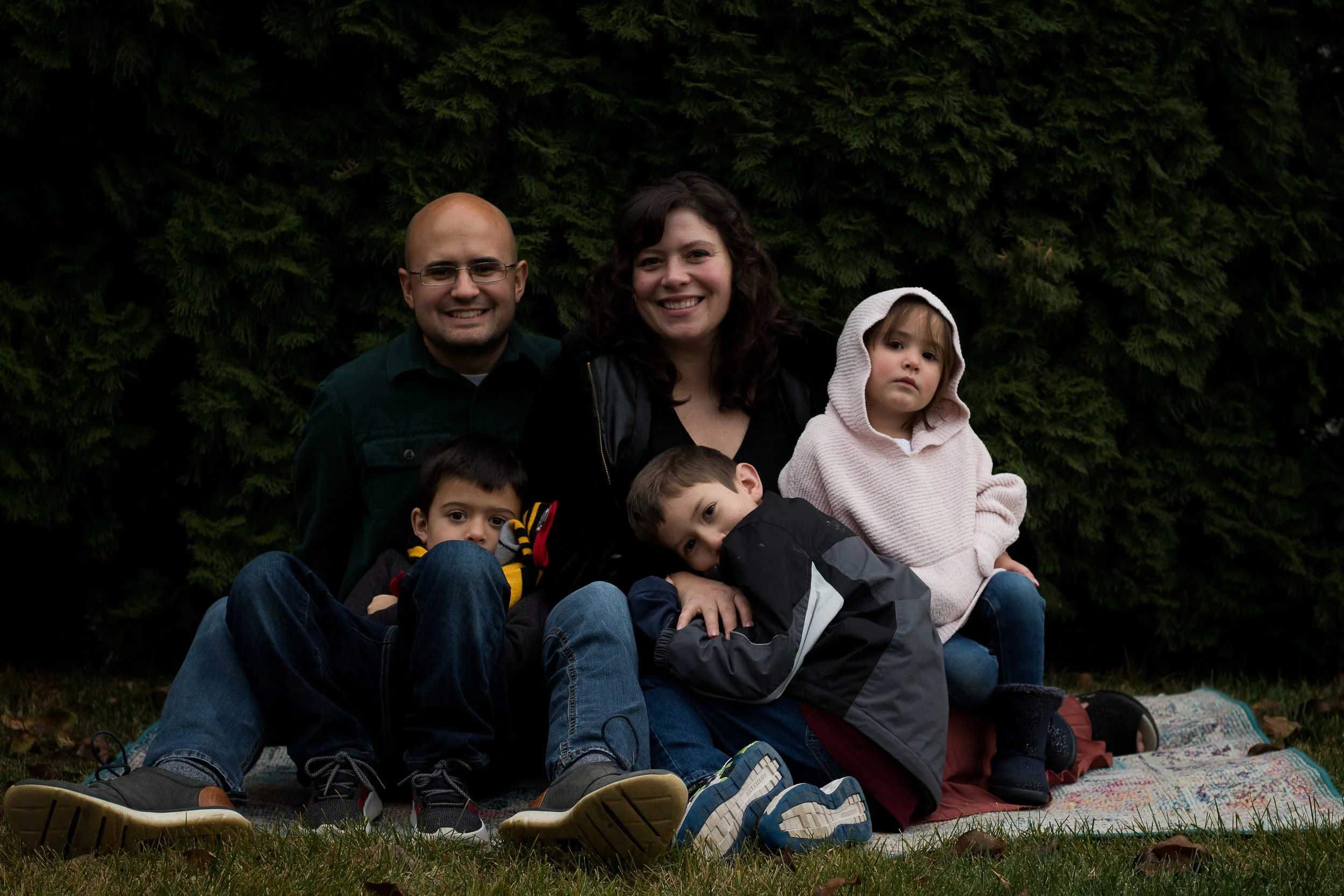 A family of five sitting on a blanket outdoors in front of evergreen trees. The family includes an adult man and woman with three children, one of whom is a young girl in a hoodie. The group is smiling and close together.