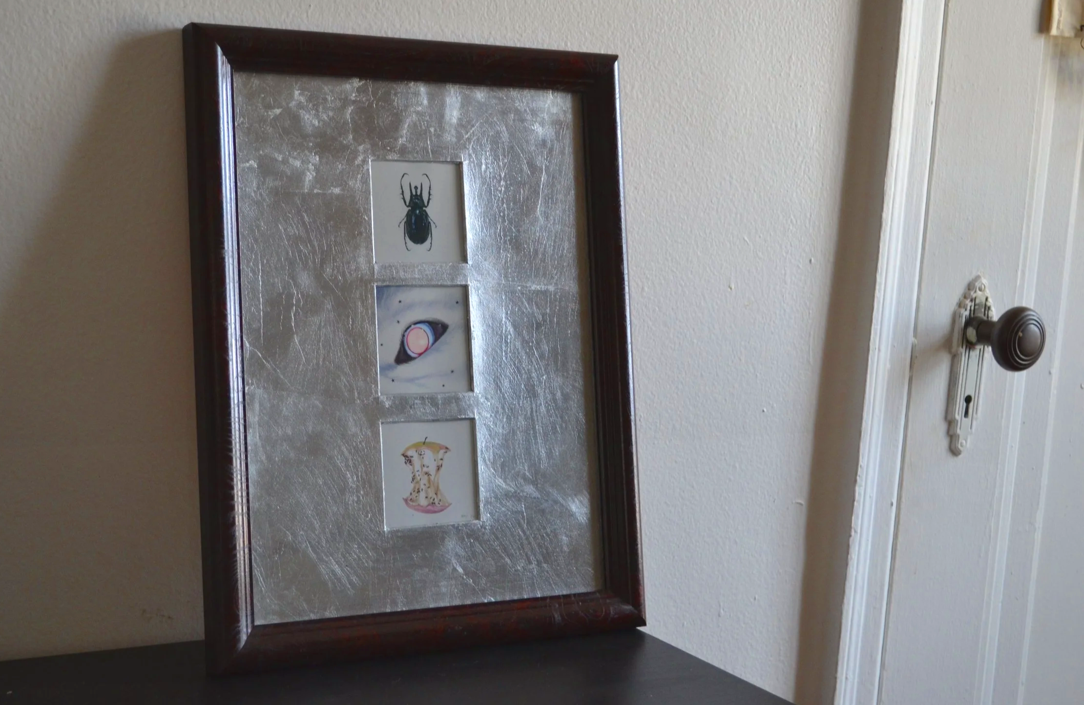 Silver-matted framed artwork with three small illustrations: a black beetle, a canine eye, and an apple core covered with ants.