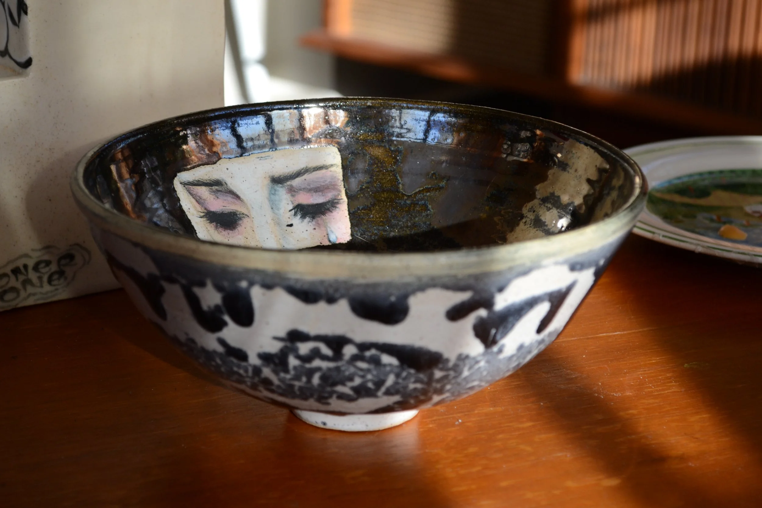 A black and white ceramic bowl with a hand-painted face of a woman with closed eyes, pink blush, long lashes, and a tear on her cheek, placed on a wooden surface.