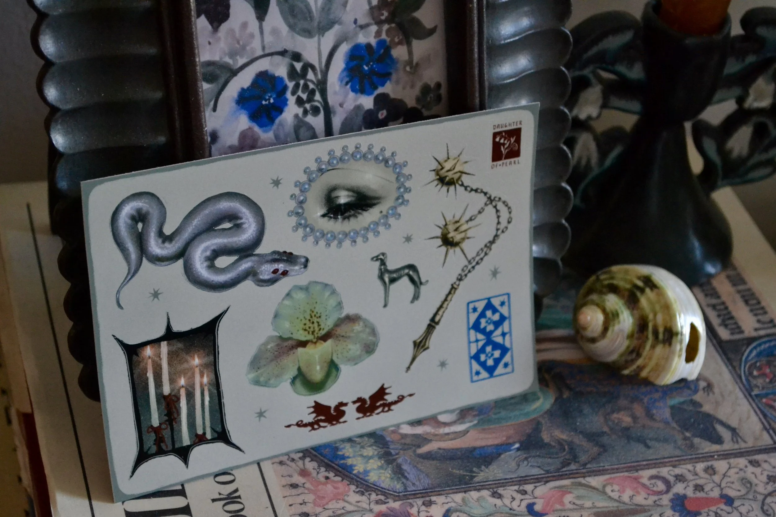 A sticker sheet with various images including a snake, eye, flower, dog, flail, candle, and dragons, placed on a surface with decorative items and a shell nearby.