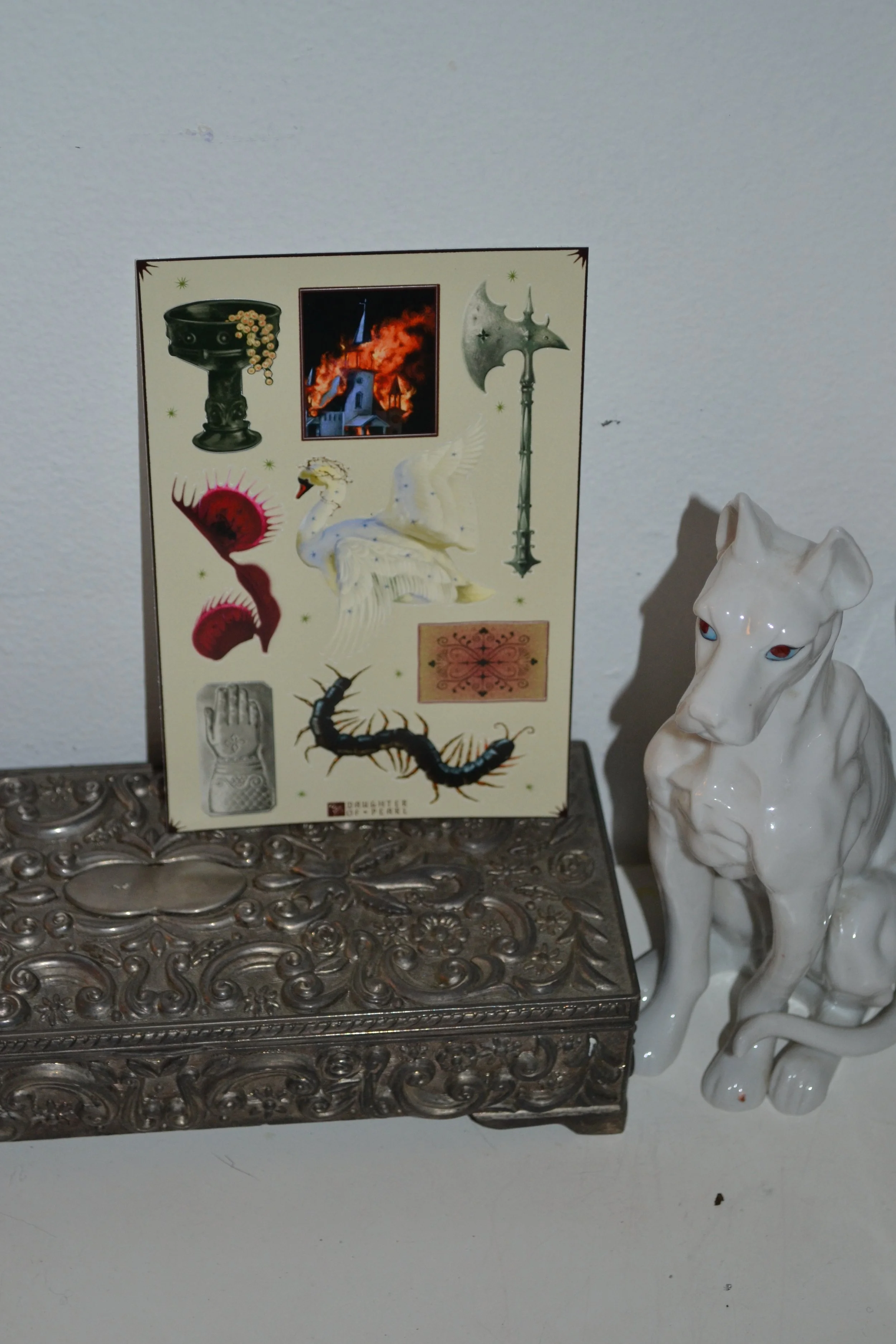 Decorative silver jewelry box, a white ceramic sculpture of a dog with red eyes, and a sheet of fantasy stickers