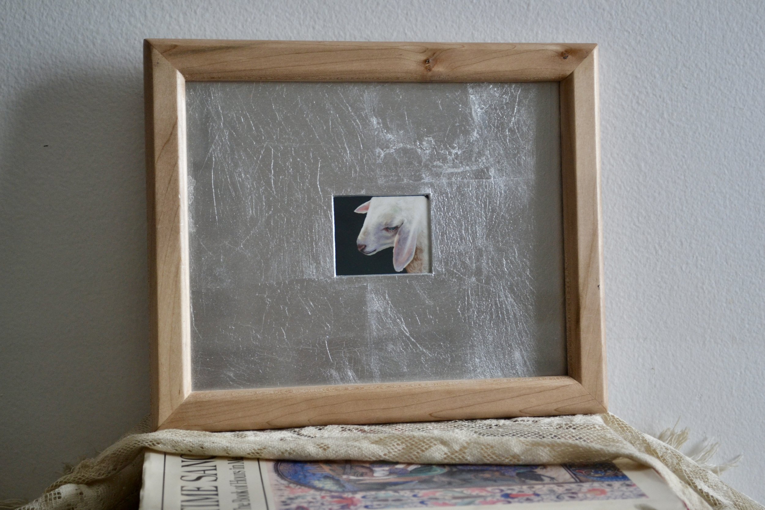 A silver-matted illustration of a lamb in a wooden frame.