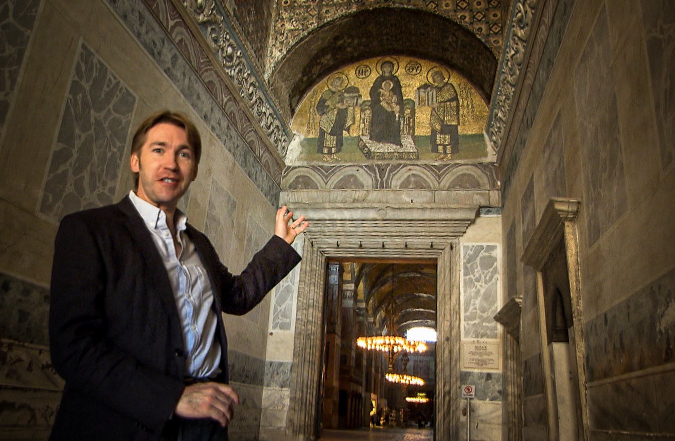 A man in a black blazer and white shirt pointing at a religious mosaic on the ceiling inside a historic building with stone walls and archways.