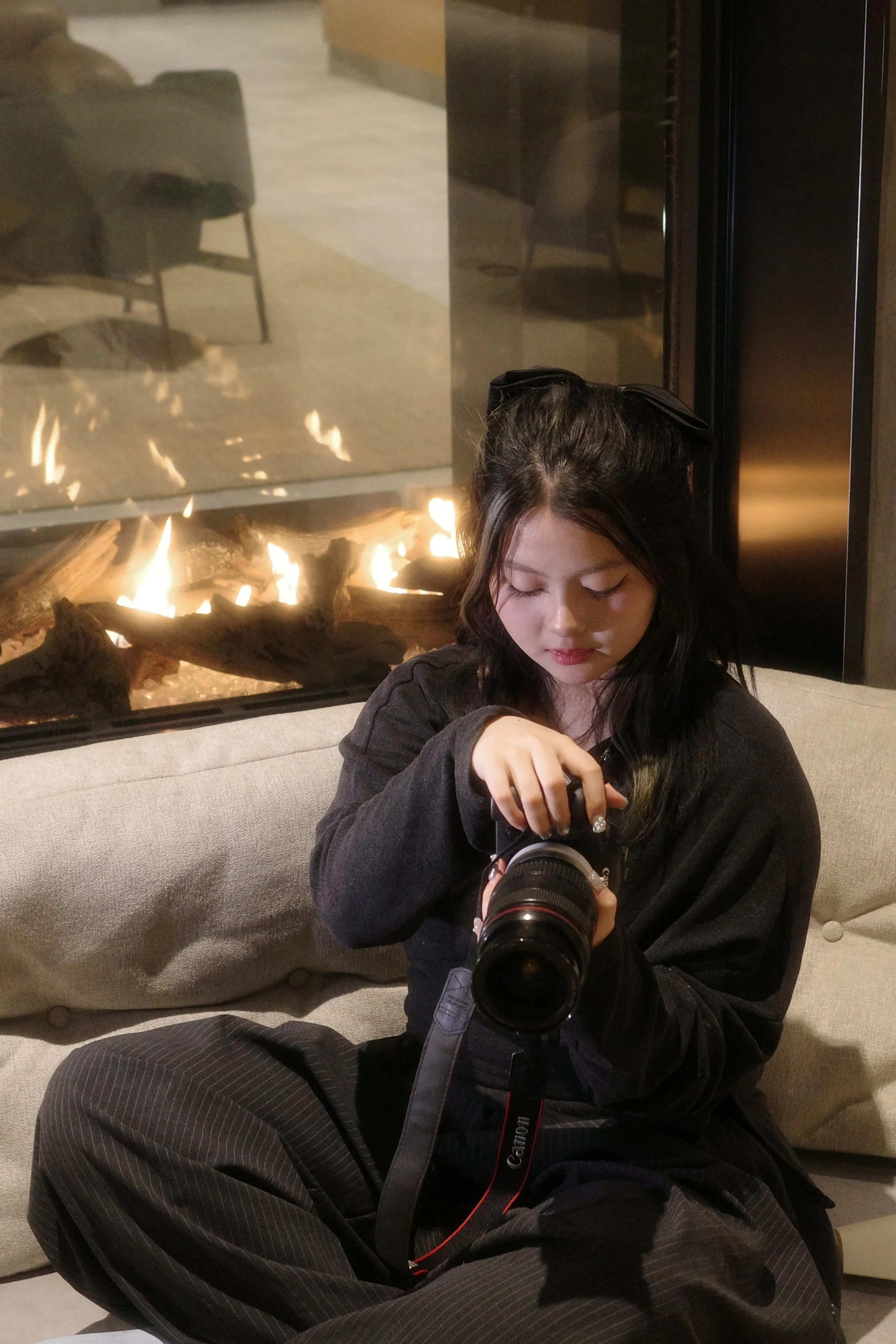 Young woman sitting on a beige sofa near a lit fireplace, holding a Canon camera and looking down at it.