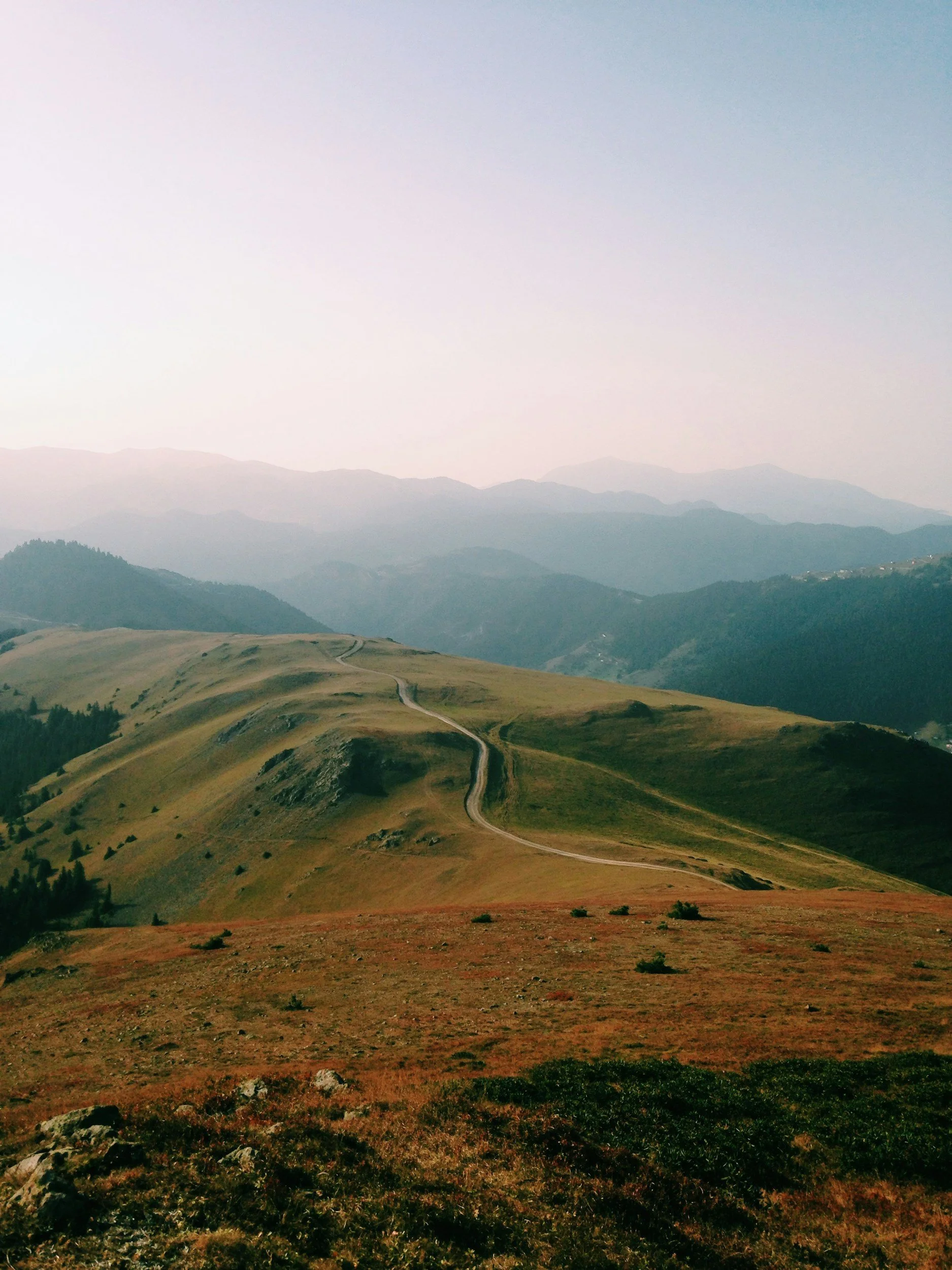 Hilly landscape with a winding dirt path, grassy terrain, and distant mountain ranges under a hazy sky.