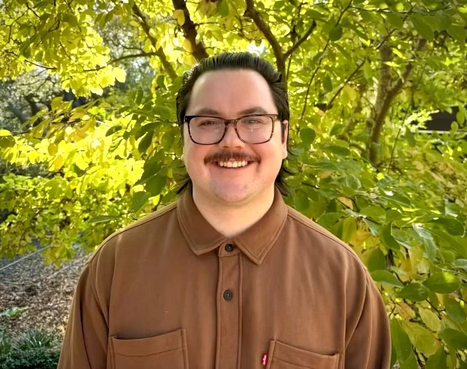 A man with glasses and a mustache smiling outdoors in front of green leafy trees.