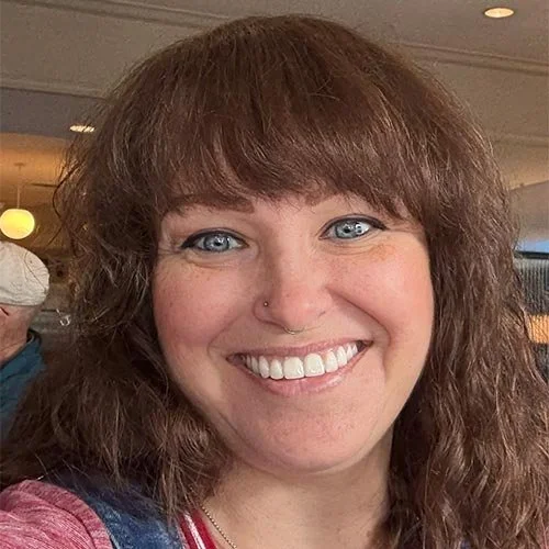 Close-up of a smiling woman with curly brown hair, blue eyes, and a nose piercing, in a public indoor setting.
