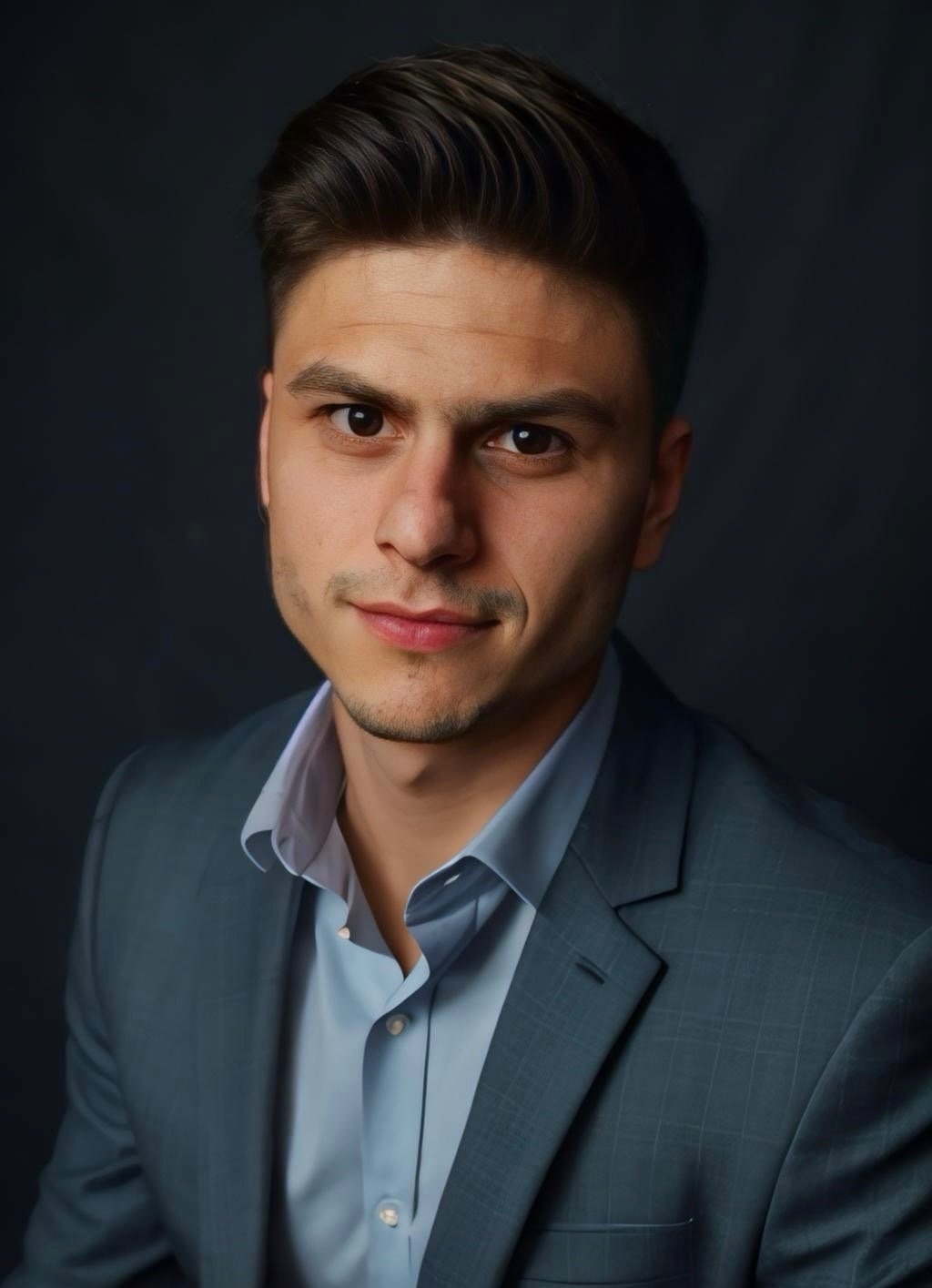A young man with dark hair, wearing a light blue dress shirt and a gray business suit, smiling at the camera against a dark background.
