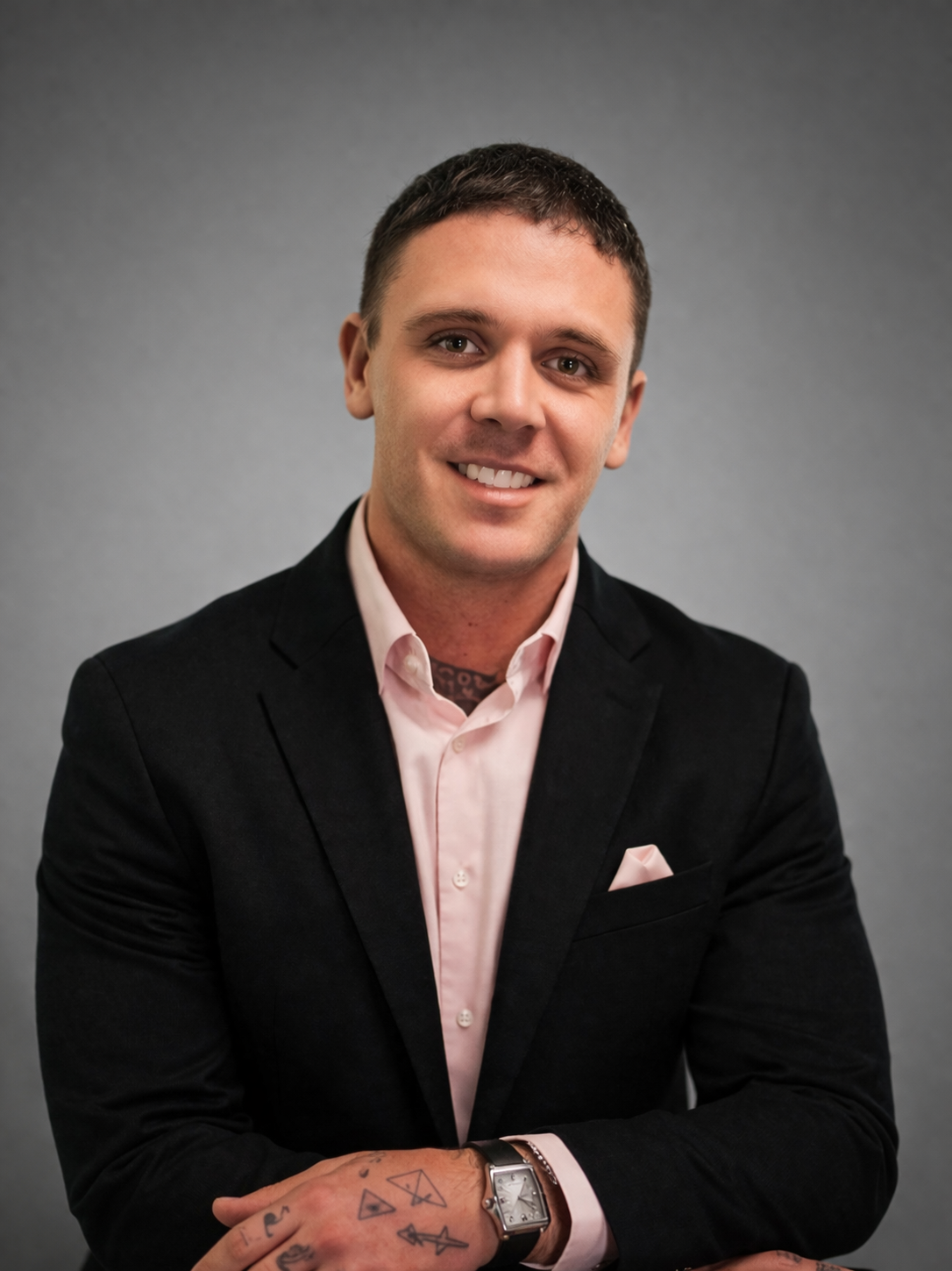 Portrait of a smiling young man with short dark hair, dressed in a black blazer, light pink shirt, and a rose gold watch, standing against a gray background.