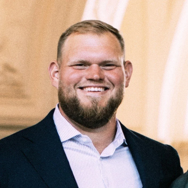 A smiling man with a beard and short hair, wearing a navy blazer and a light-colored dress shirt, standing indoors.
