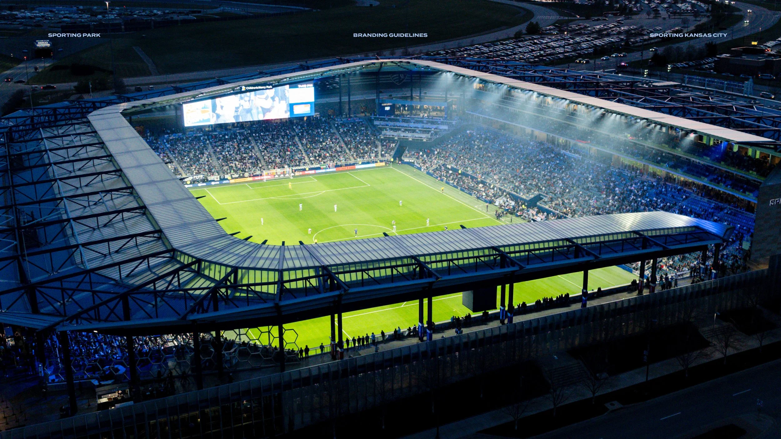 An aerial view of a illuminated soccer stadium at dusk, filled with spectators, with players on the field.
