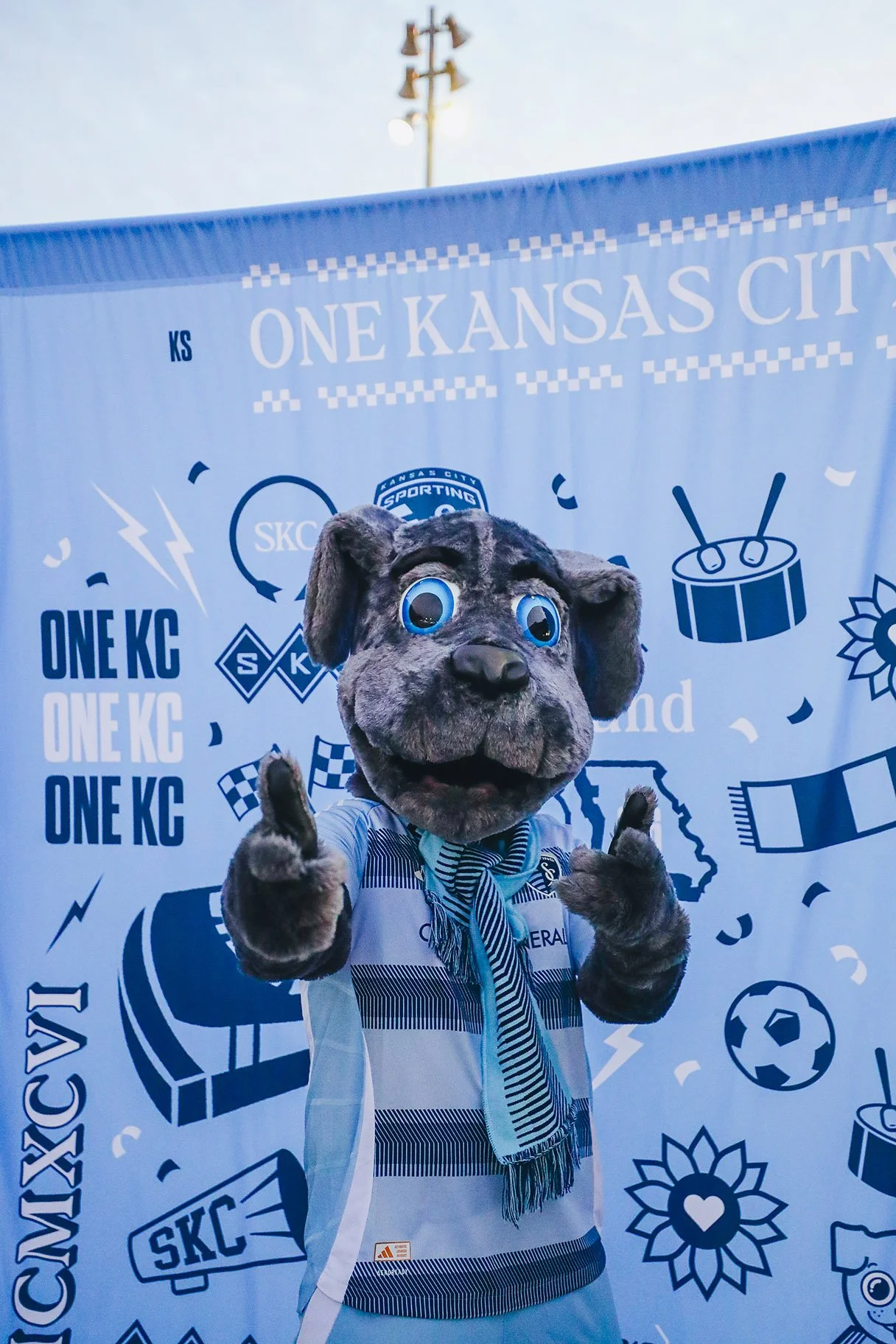 Mascot in a dog costume giving a thumbs up at a sporting event with a blue and white banner in the background that says 'One Kansas City.'