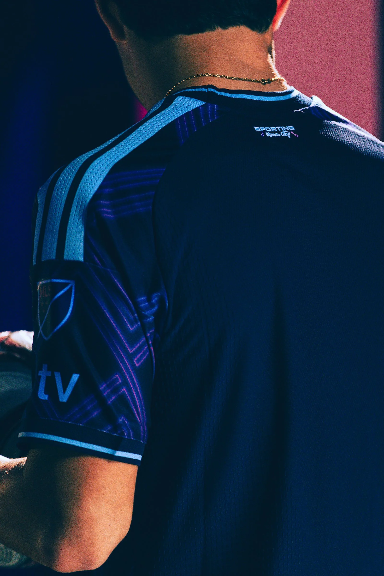 Back view of a person wearing a dark blue sports jersey with purple and light blue accents, and a gold chain necklace, with the word 'SPORTING' visible on the upper back.