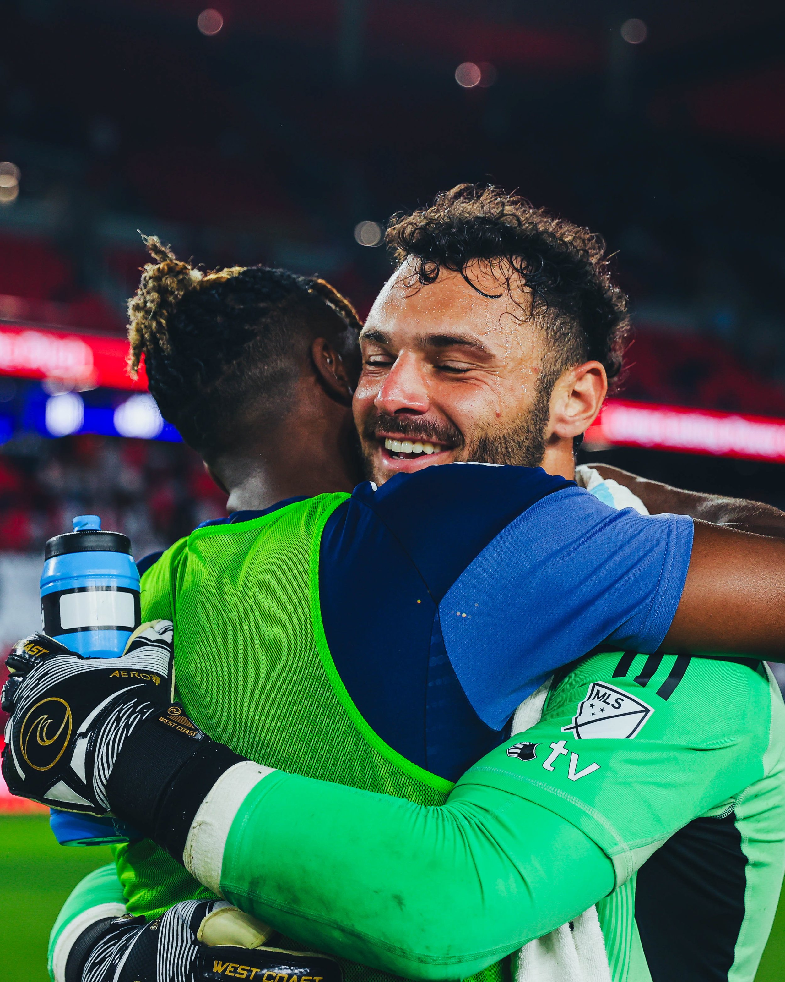 Two soccer players hugging after a match, one in a bright green goalkeeper jersey and the other in a blue jersey, on a stadium field.
