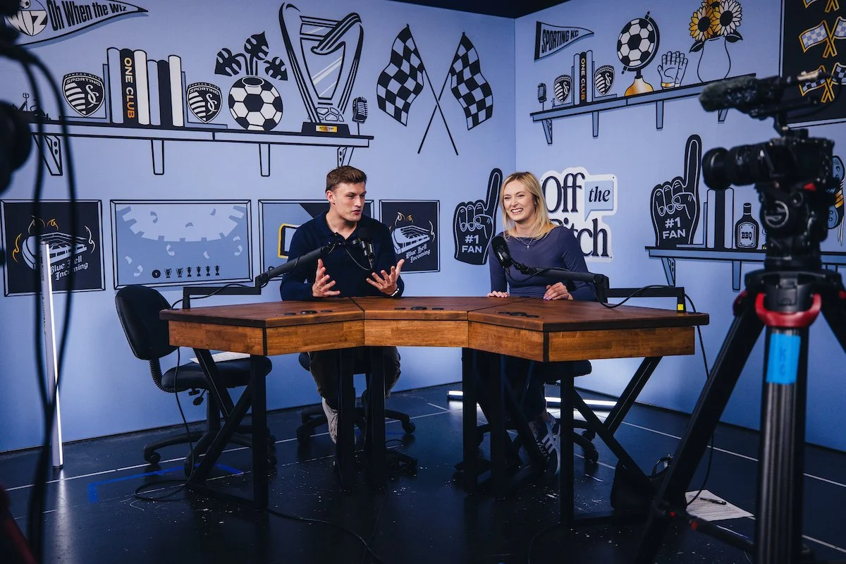 Two people, a man and a woman, sitting at a wooden table with microphones in front of them, recording a podcast or interview in a room decorated with sports and soccer-themed illustrations and memorabilia on the walls.