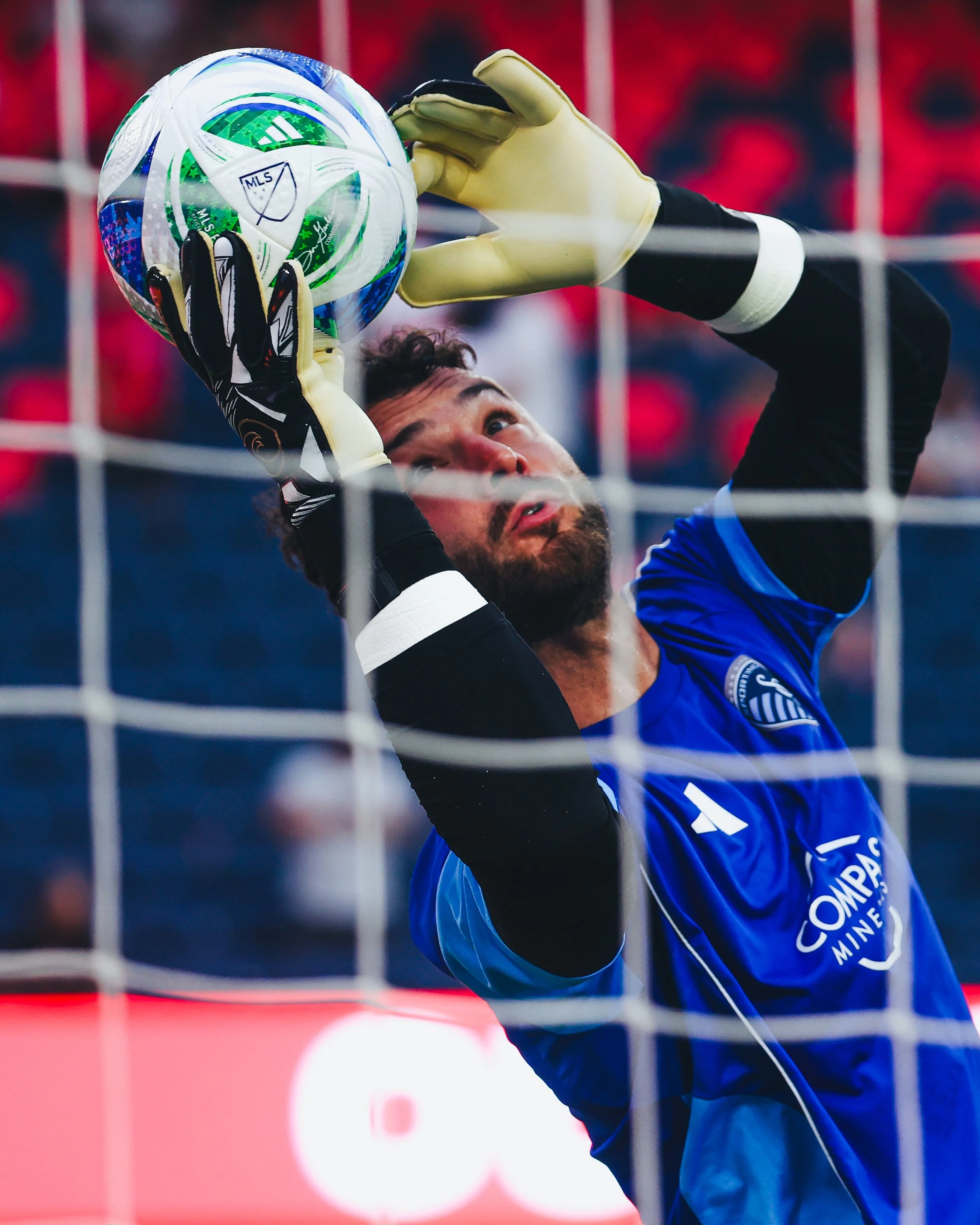A soccer player in a blue jersey and black gloves reaching to catch a green and white MLS-branded soccer ball near the goal net.