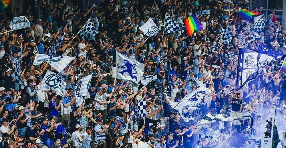 A large crowd of sports fans in stadium, many waving flags and wearing team colors, cheering and celebrating.