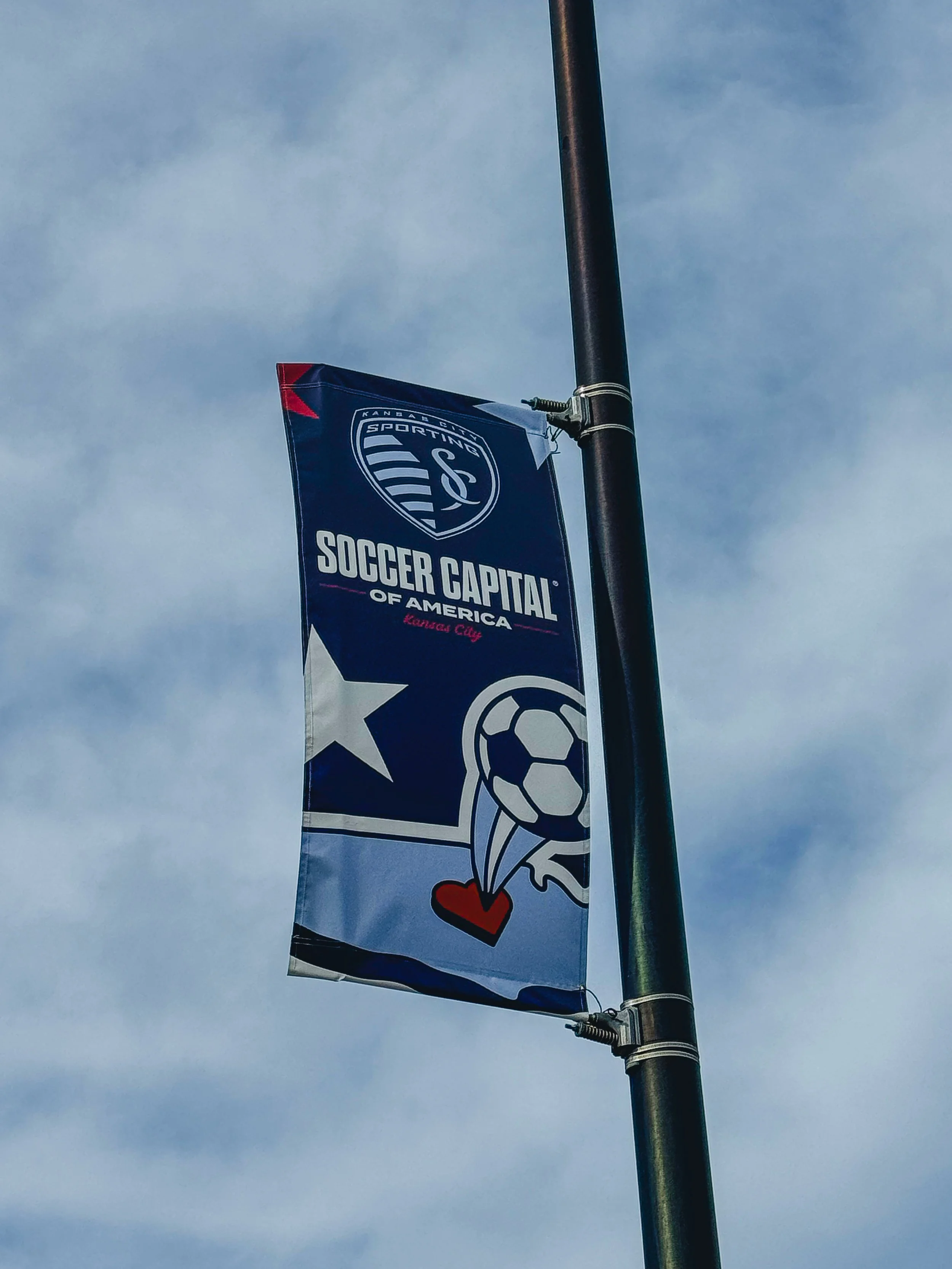 Flag with Kansas City Sporting Soccer logo, a soccer ball with steam and a heart, and the text 'Soccer Capital of America' and 'Kansas City' on it, attached to a black flagpole against a cloudy sky.