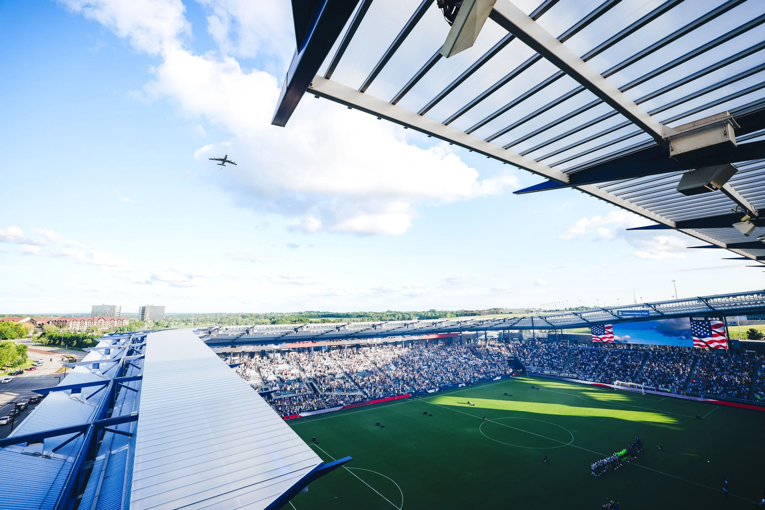 A view of a soccer stadium filled with spectators, with a large digital screen displaying flags and an American flag in the stands, under a partly cloudy sky with an airplane flying overhead.