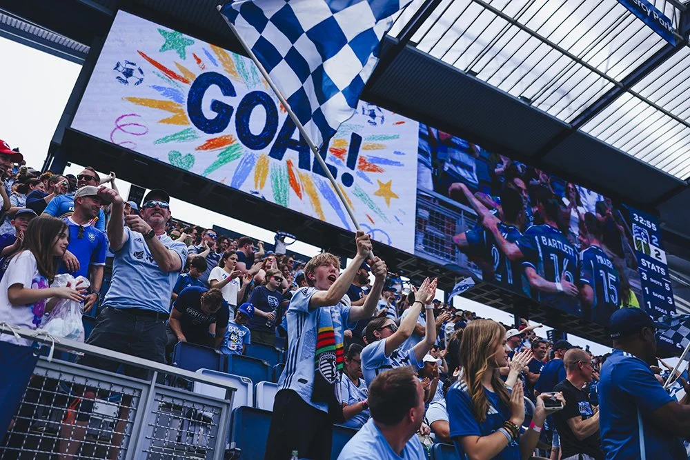 Soccer fans celebrating with a large digital scoreboard displaying a colorful drawing of a goal and a cheering crowd.