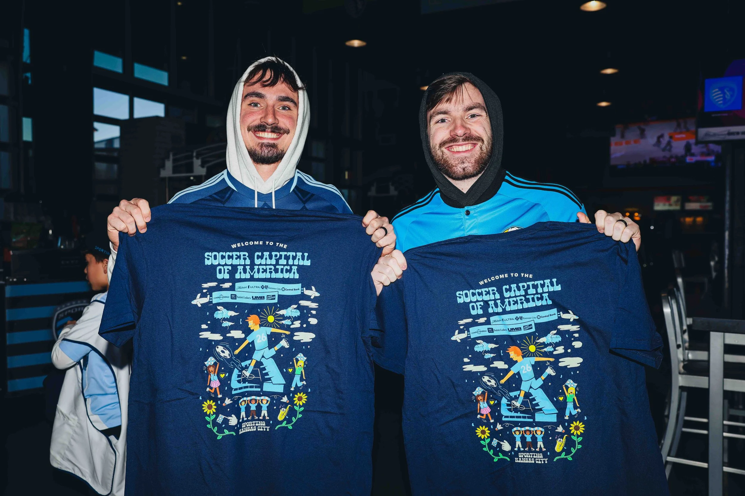 Two smiling men holding blue T-shirts with colorful graphics and the text 'Welcome to the Soccer Capital of America' in an indoor sports venue.