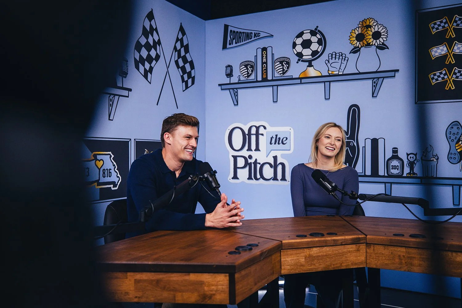 Two people, a man and a woman, sitting at a table with microphones during a podcast recording. The background features a mural with sports-themed illustrations and the text 'Off the Pitch'.