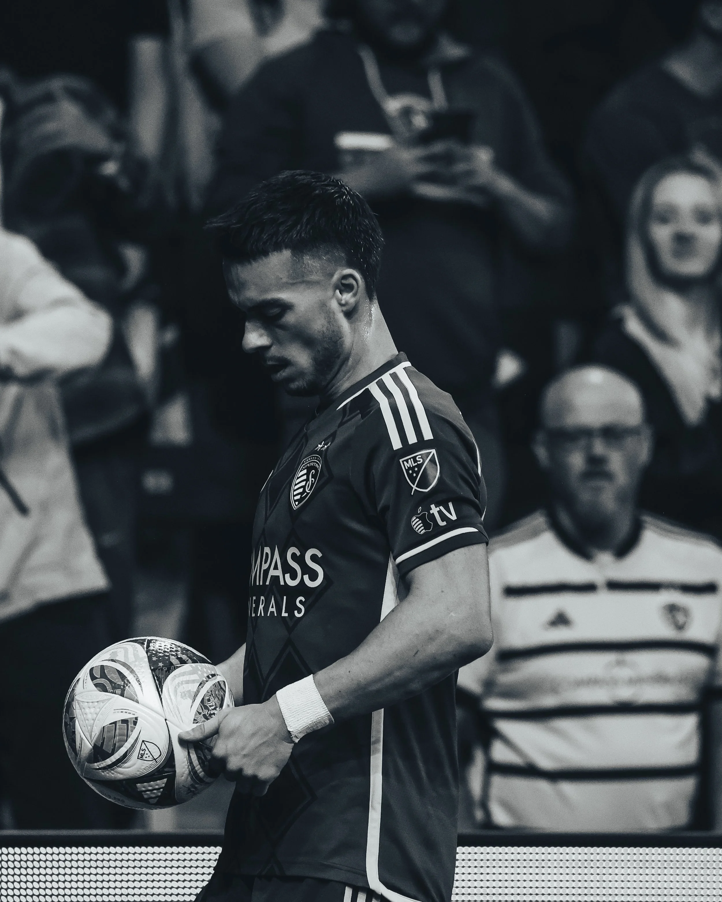 A soccer player holding a ball on the field, wearing a dark jersey with sponsor logos, with spectators in the background.