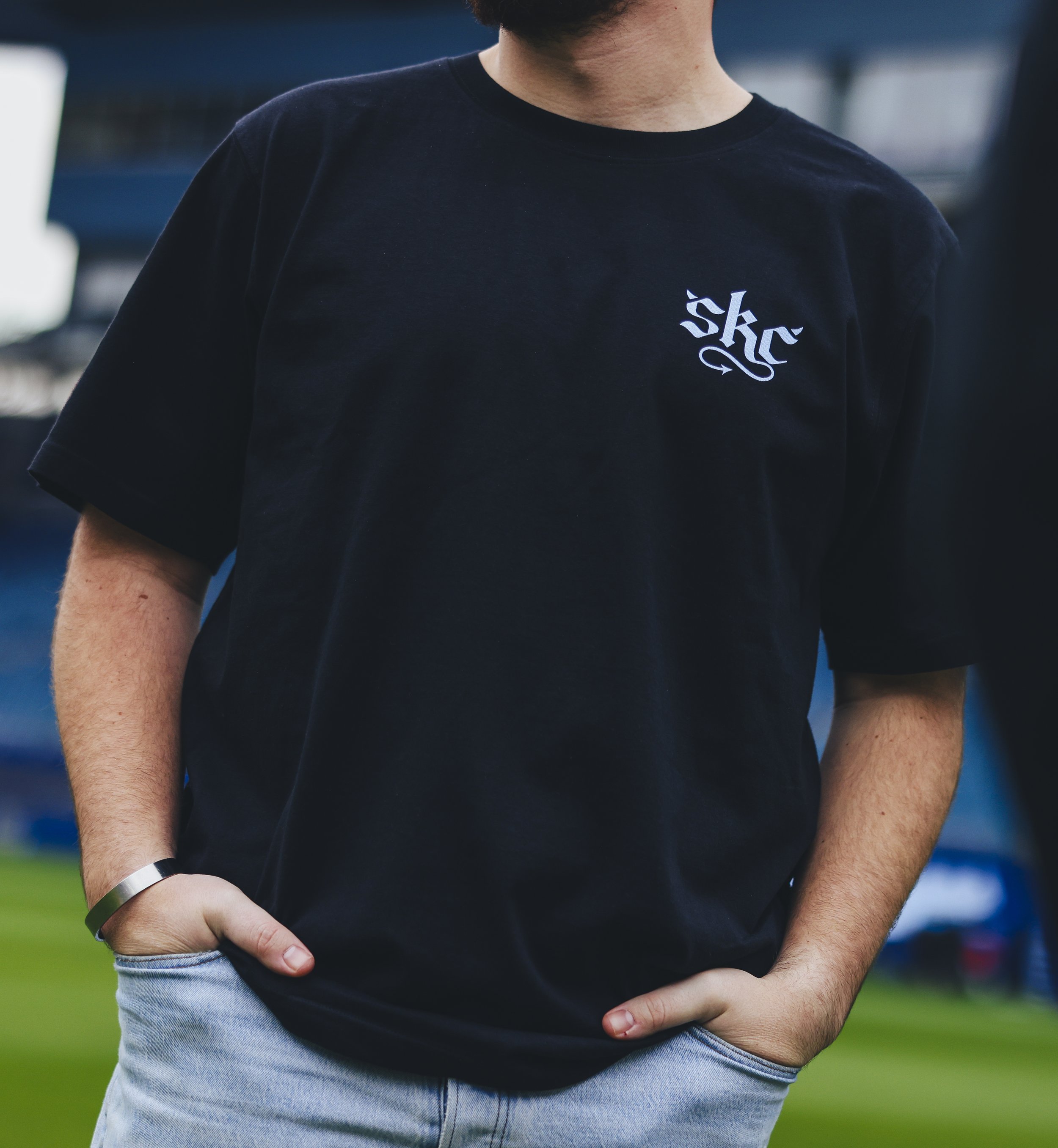 Close-up of a person wearing a black T-shirt with 'skc' logo on the chest, standing outdoors with hands in their pockets.