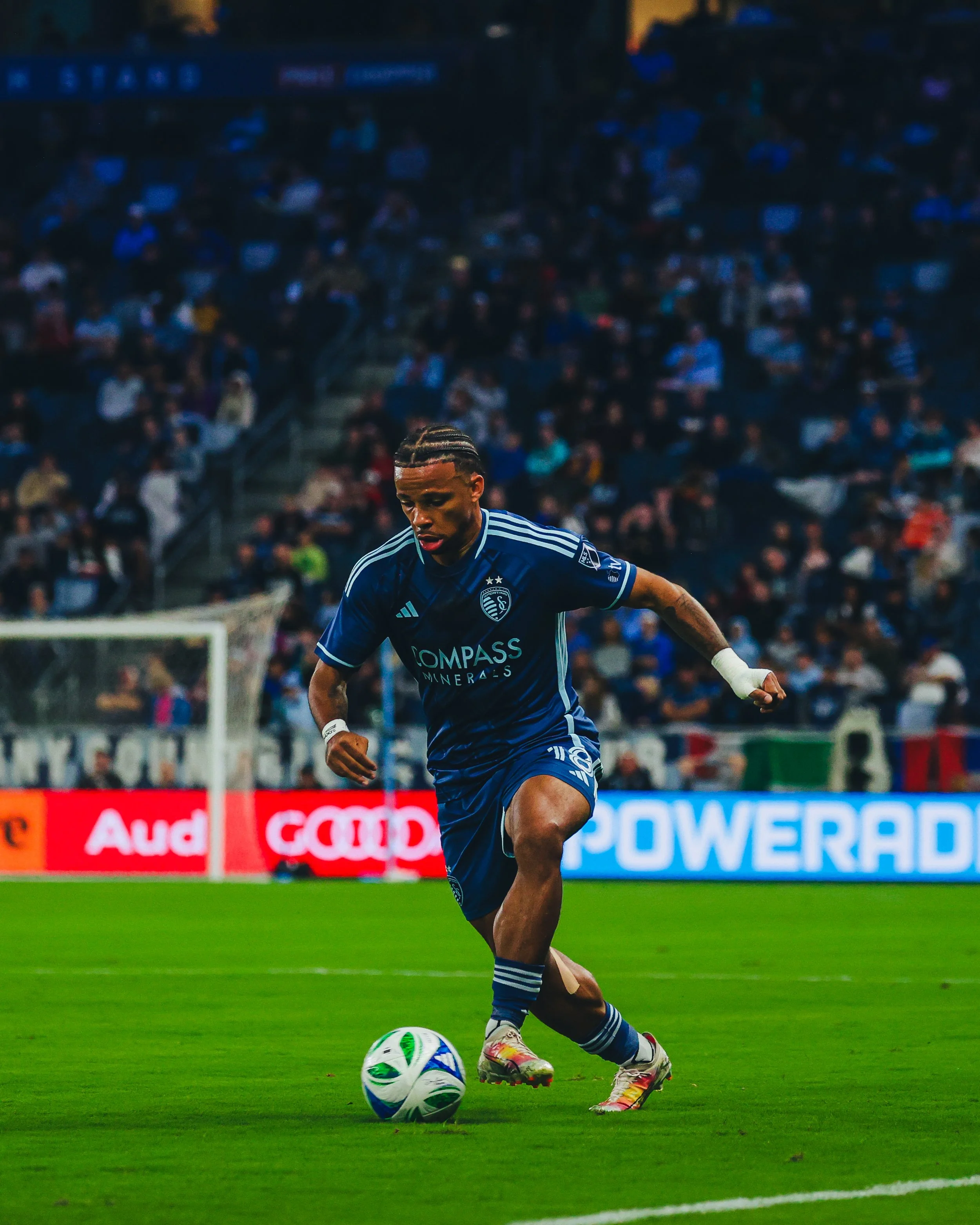 A soccer player in a blue jersey is running with a soccer ball on the field during a game, with a crowd of spectators in the background.