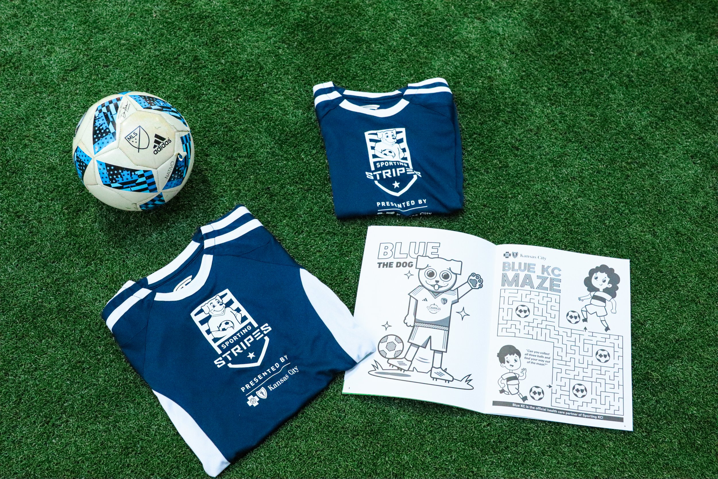 Soccer ball, two folded blue Kansas City Sporting Shirts with white logos, and a black and white coloring activity book on artificial green turf.