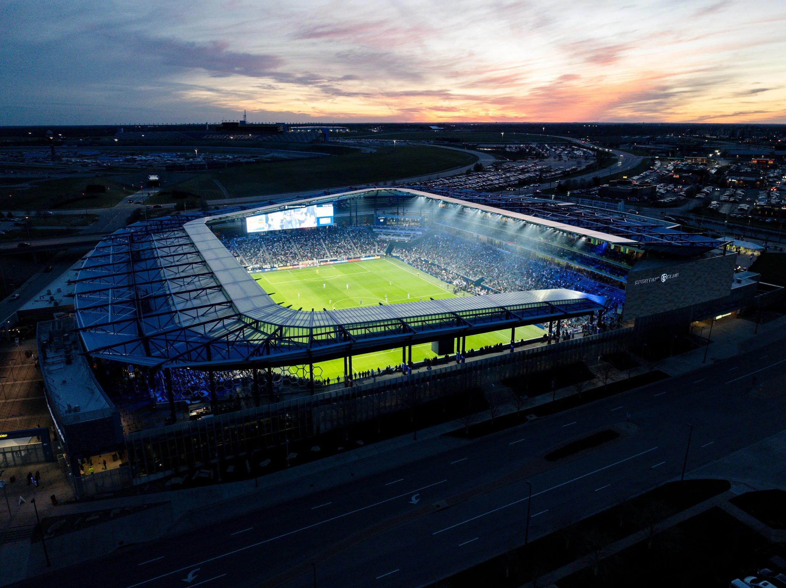 Aerial view of a soccer stadium filled with spectators at dusk, with a sunset sky in the background.