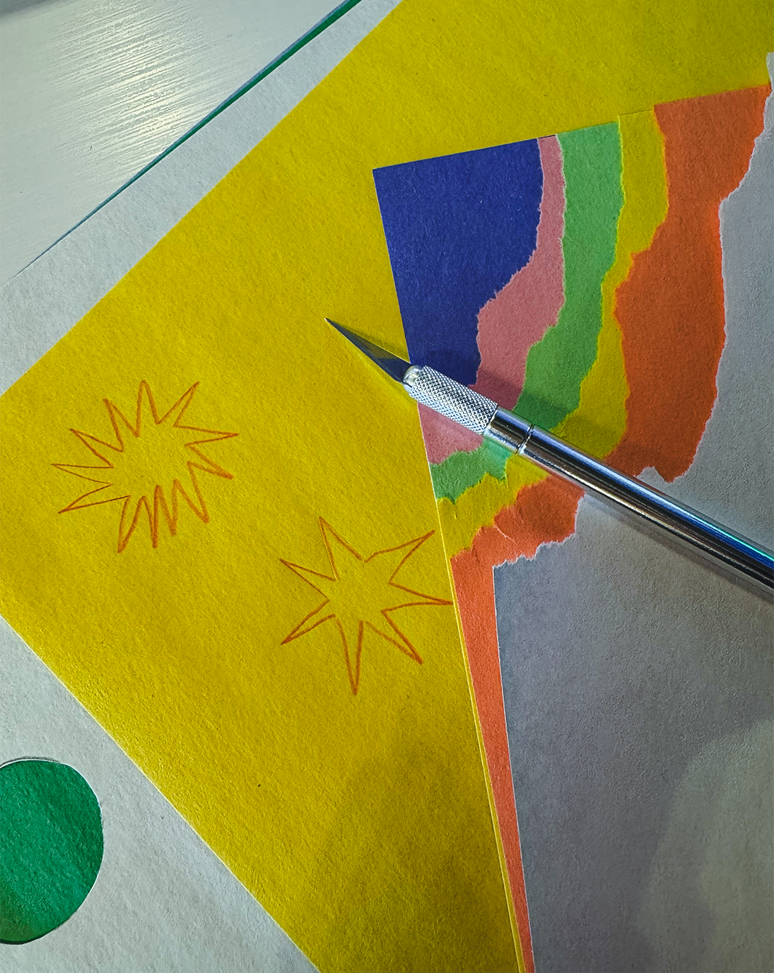 Close-up of a craft project with colored paper, a craft knife, and two star-shaped sketches on yellow paper.