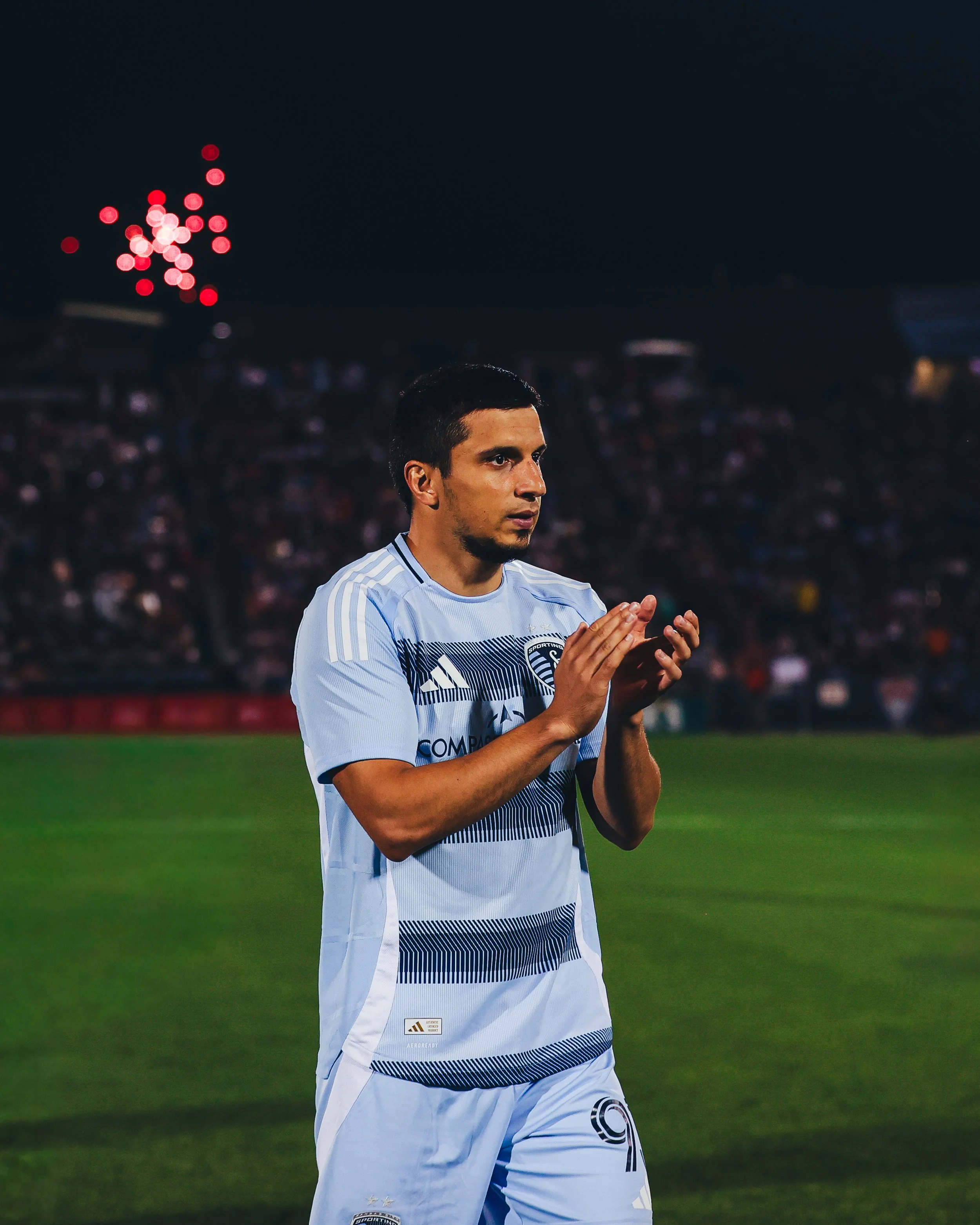 A male soccer player in a light blue and navy uniform claps his hands during a match, with a blurred crowd and stadium lights in the background.