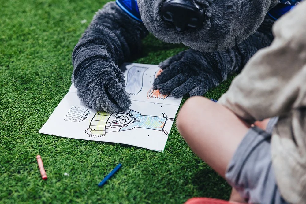 A dog and a child sit on artificial grass, coloring a printable picture of a dog with crayons.