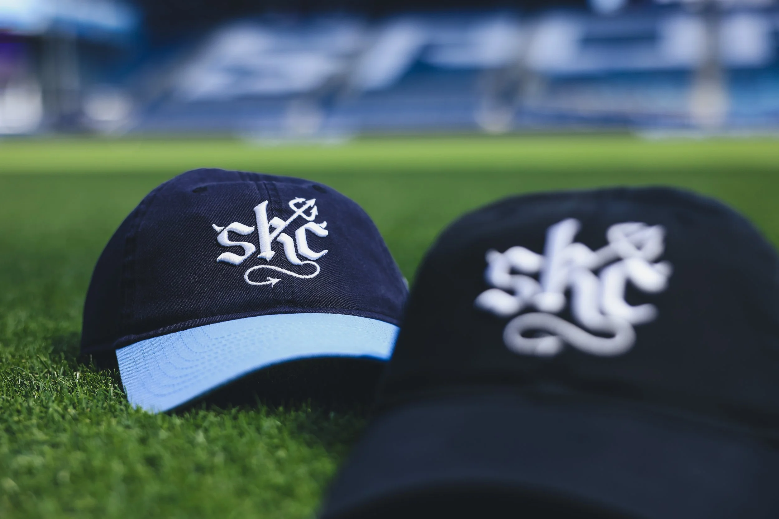 Two black and blue baseball caps with a white logo placed on a grass field, with a stadium blurred in the background.