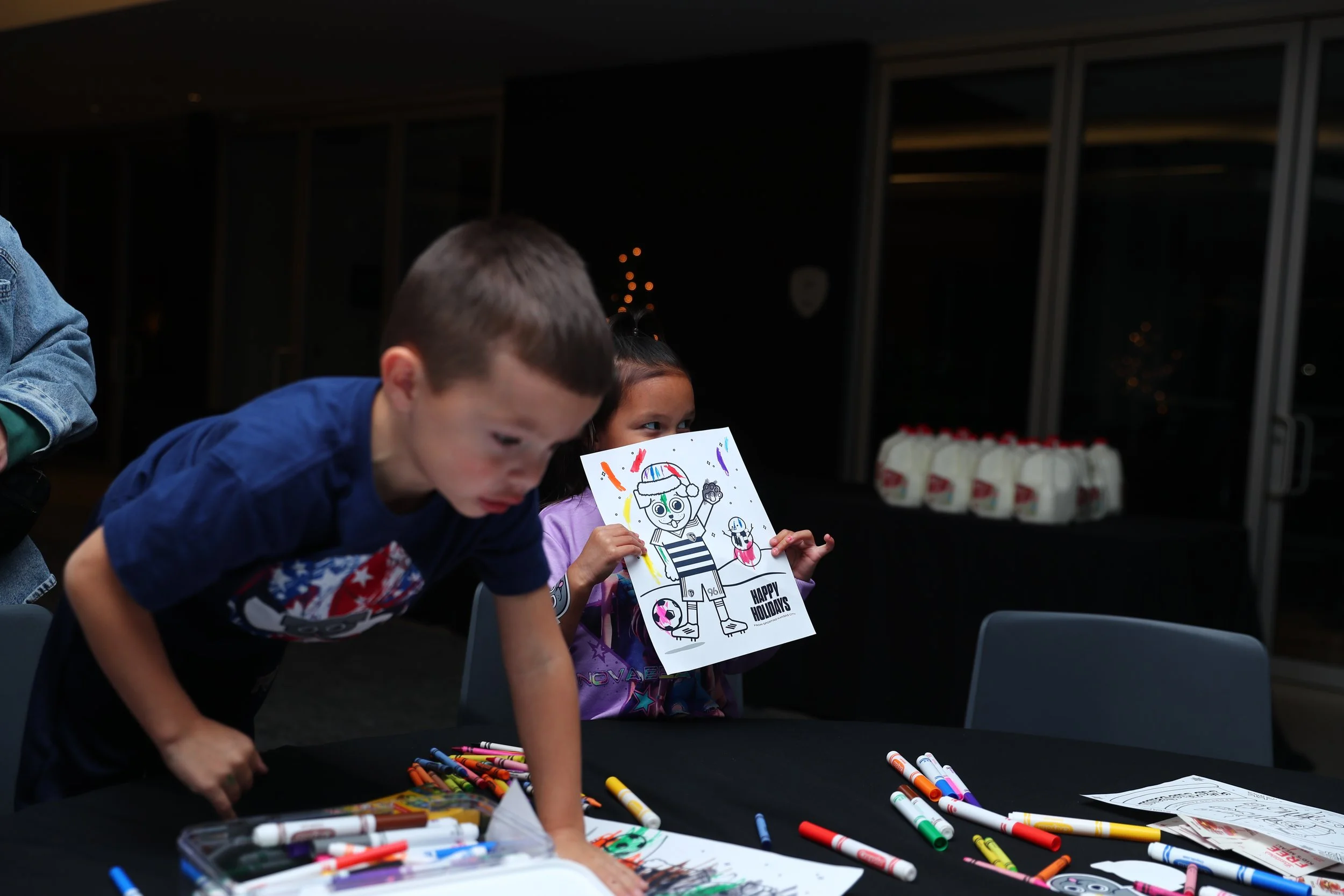 Two children at a birthday party, one girl holding a coloring page with a holiday and winter theme, the boy leaning over the table covered with Crayola crayons, with milk jugs in the background.