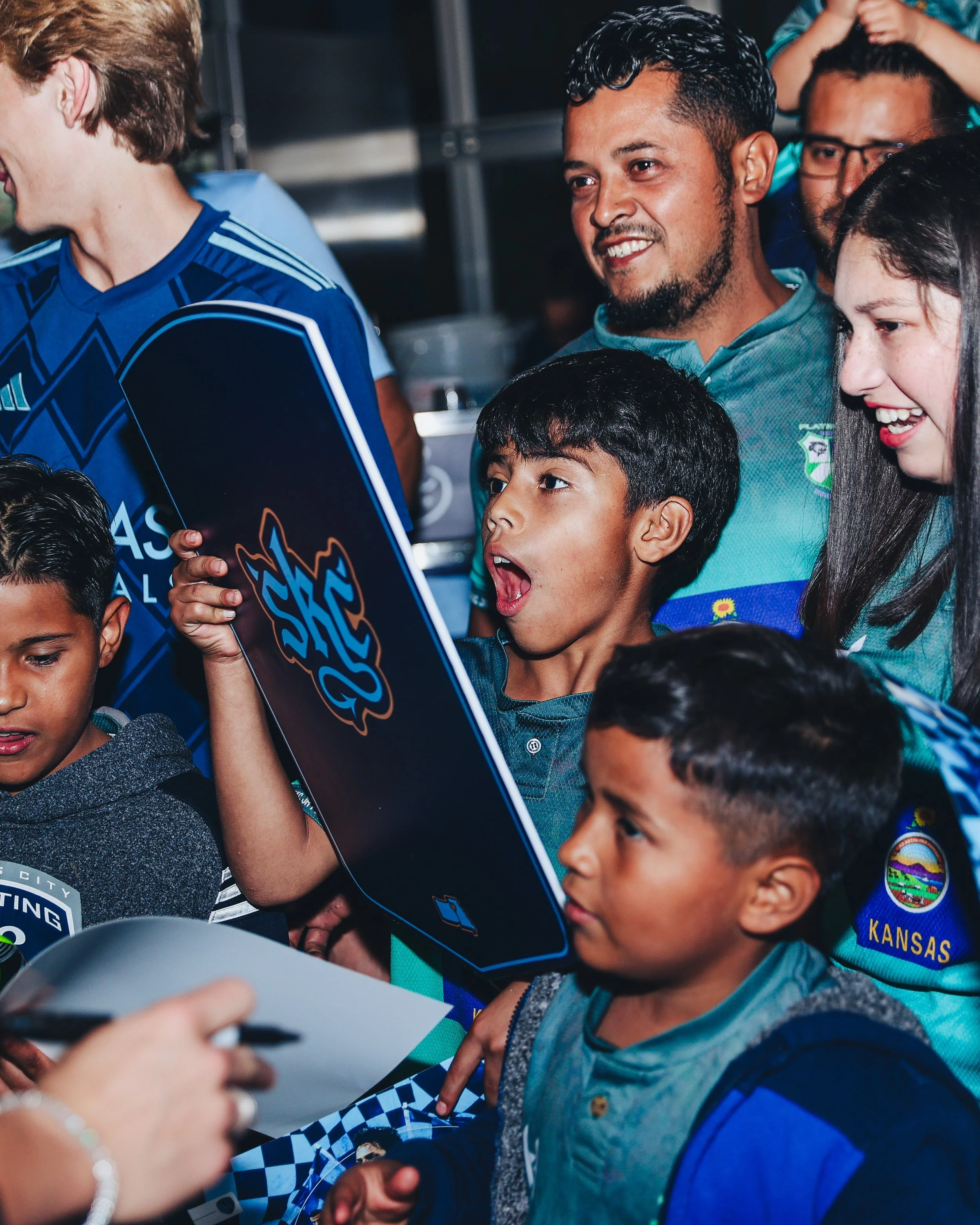 Group of children and adults gathered around a person signing an item, likely at a sports event, with excitement and joy, one boy holding a skateboard with a colorful logo.