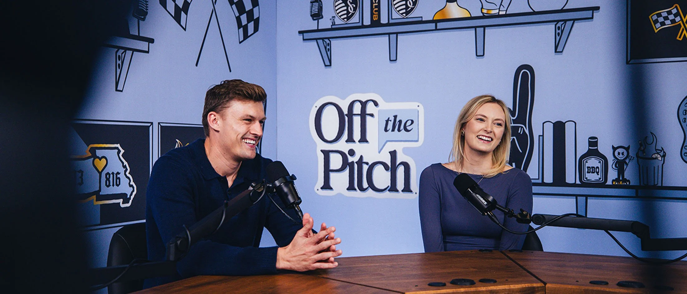Two people, a man and a woman, sitting at a table with microphones in front of them, smiling and talking during a podcast or radio show. The background features a colorful wall with illustrations of various objects and the text "Off the Pitch".