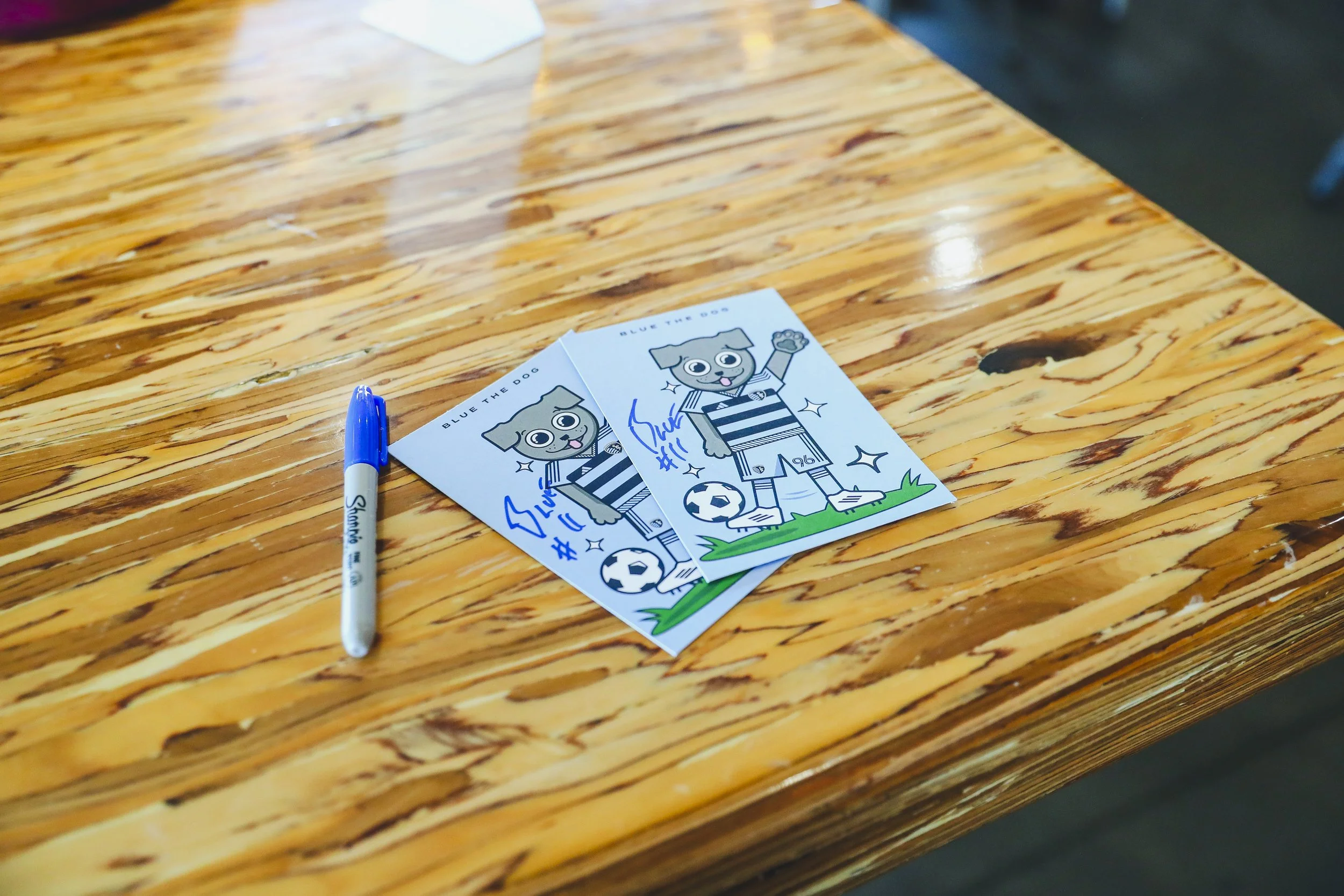 Two signed drawings of a cartoon dog dressed in a soccer uniform on a wooden table with a blue Sharpie pen nearby.