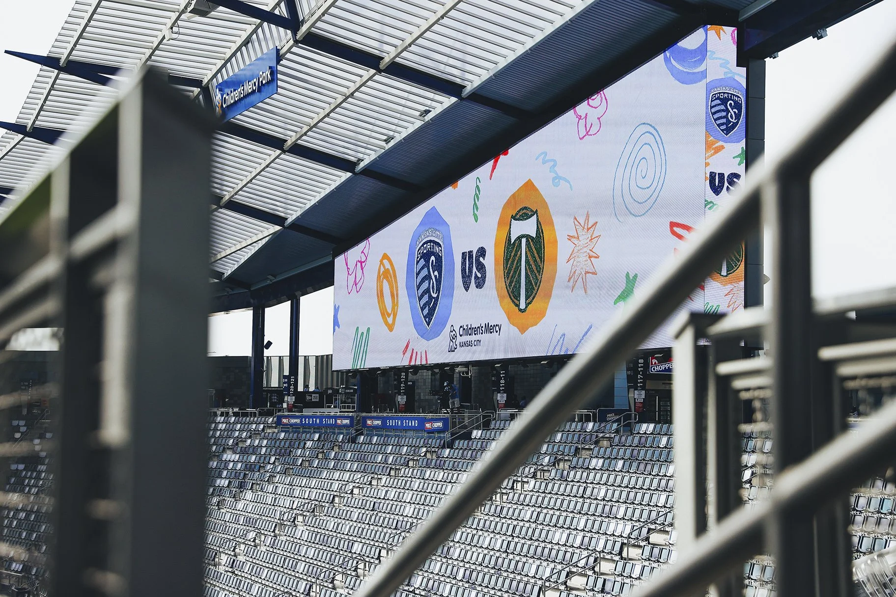 Empty stadium seats with a large LED display showing a match between Sporting Kansas City and Portland Timbers, with Children's Mercy Kansas City logo, during daytime.
