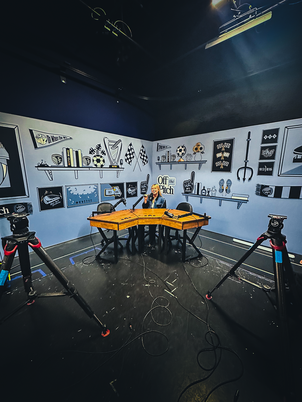 A woman sitting at a wooden table in a room with sports-themed black and white wall graphics, microphones, and camera equipment recording or broadcasting a show.