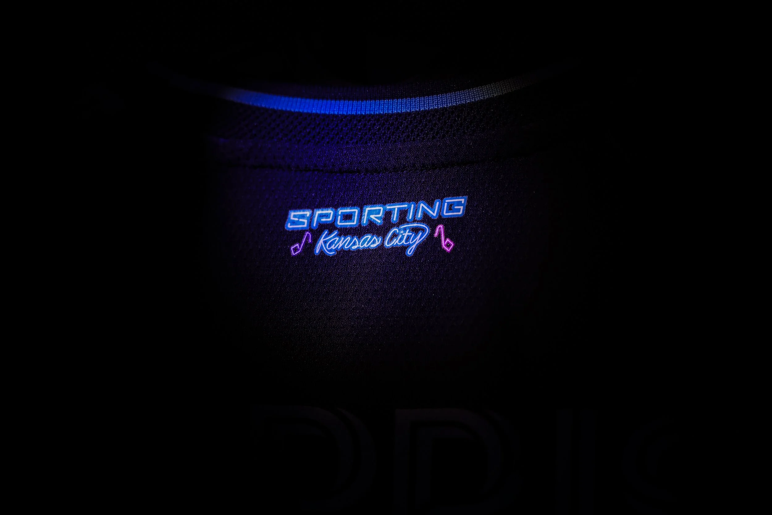 Close-up of a dark sports jersey with illuminated text reading 'SPORTING Kansas City' in neon blue and purple colors, with musical note icons.