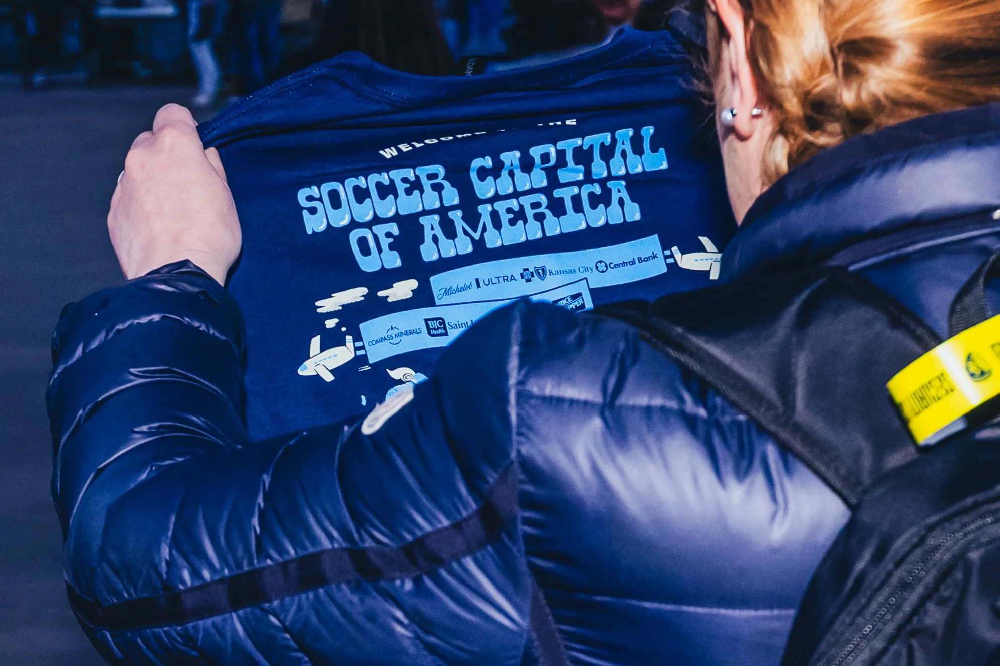 Back of a person's shirt with the text 'Soccer Capital of America' and logos of sponsors, being held by another person in a jacket, in an outdoor setting with several people in the background.