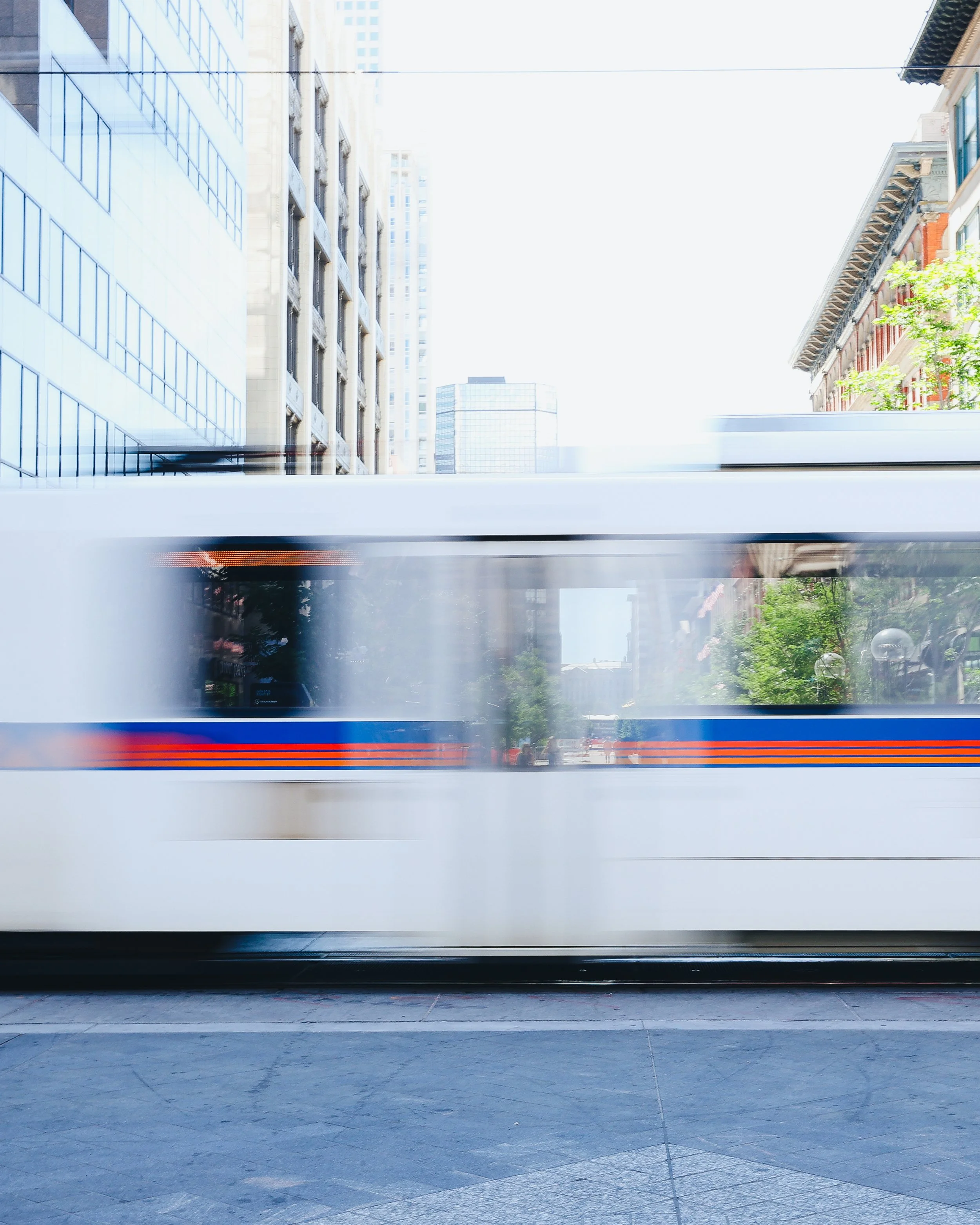 A moving white city transit train passing through an urban area with tall buildings, green trees, and a clear sky.