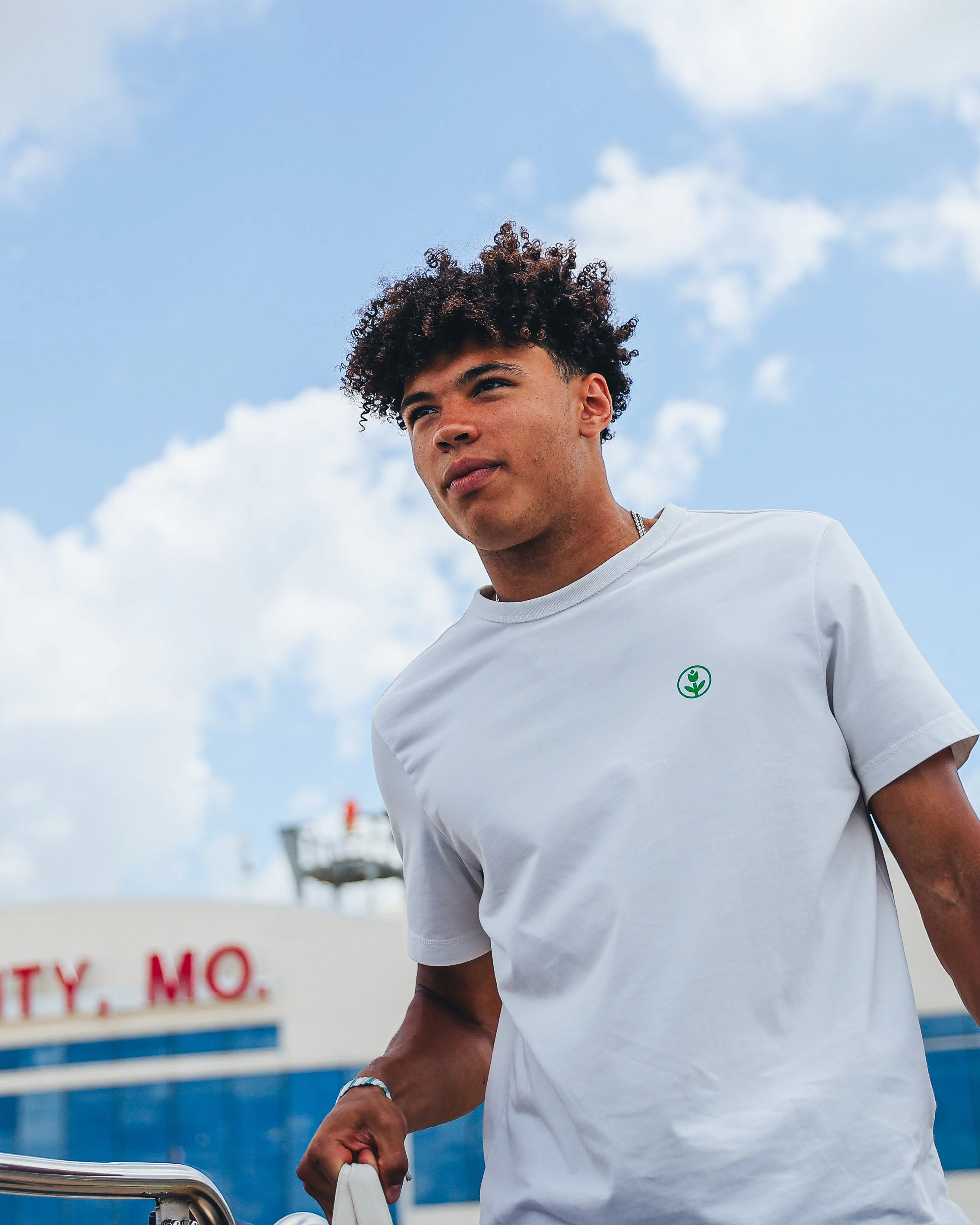 A young man with curly hair wearing a white T-shirt with a small green logo, carrying a bag, standing outdoors under a blue sky with scattered clouds.